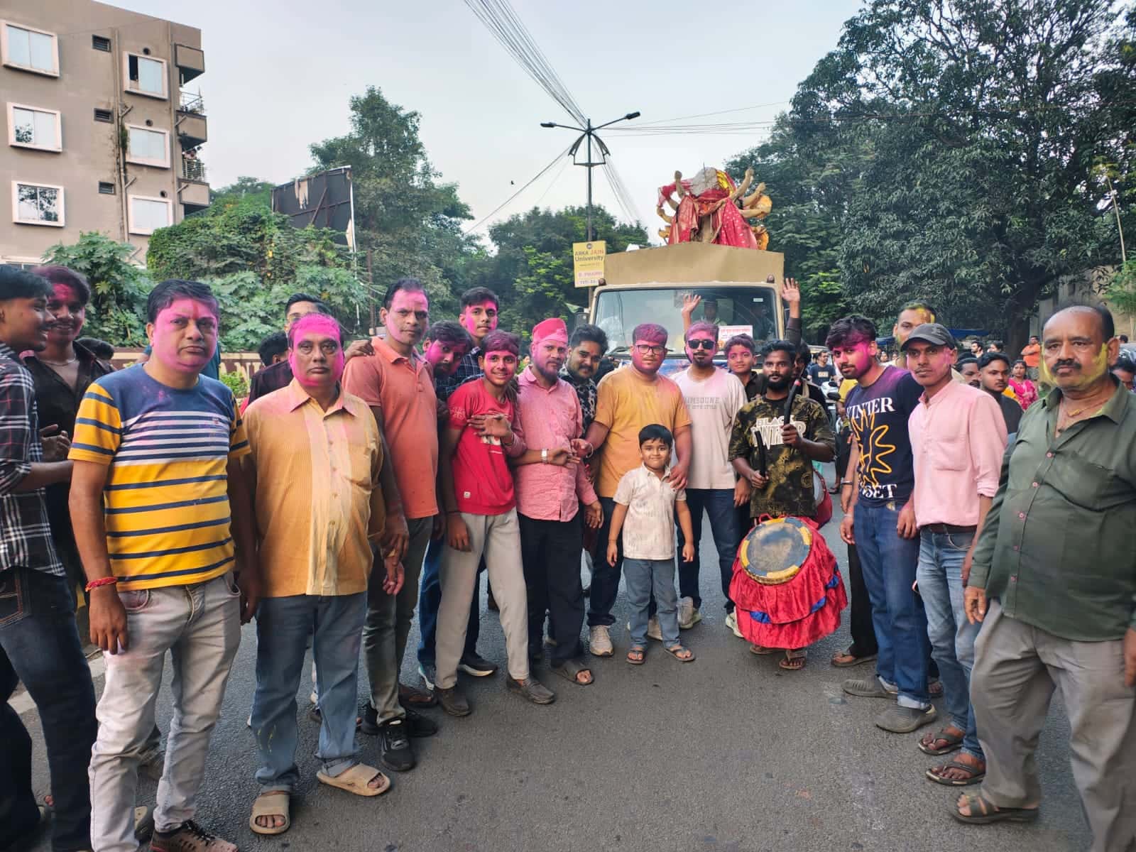 सड़क पर गुलाबी और पीले रंग से रंगे चेहरों वाले कई लोग समूह में खड़े हैं। पीछे एक ट्रक पर देवी की प्रतिमा रखी दिखाई दे रही है और एक व्यक्ति ढोल के साथ खड़ा है।