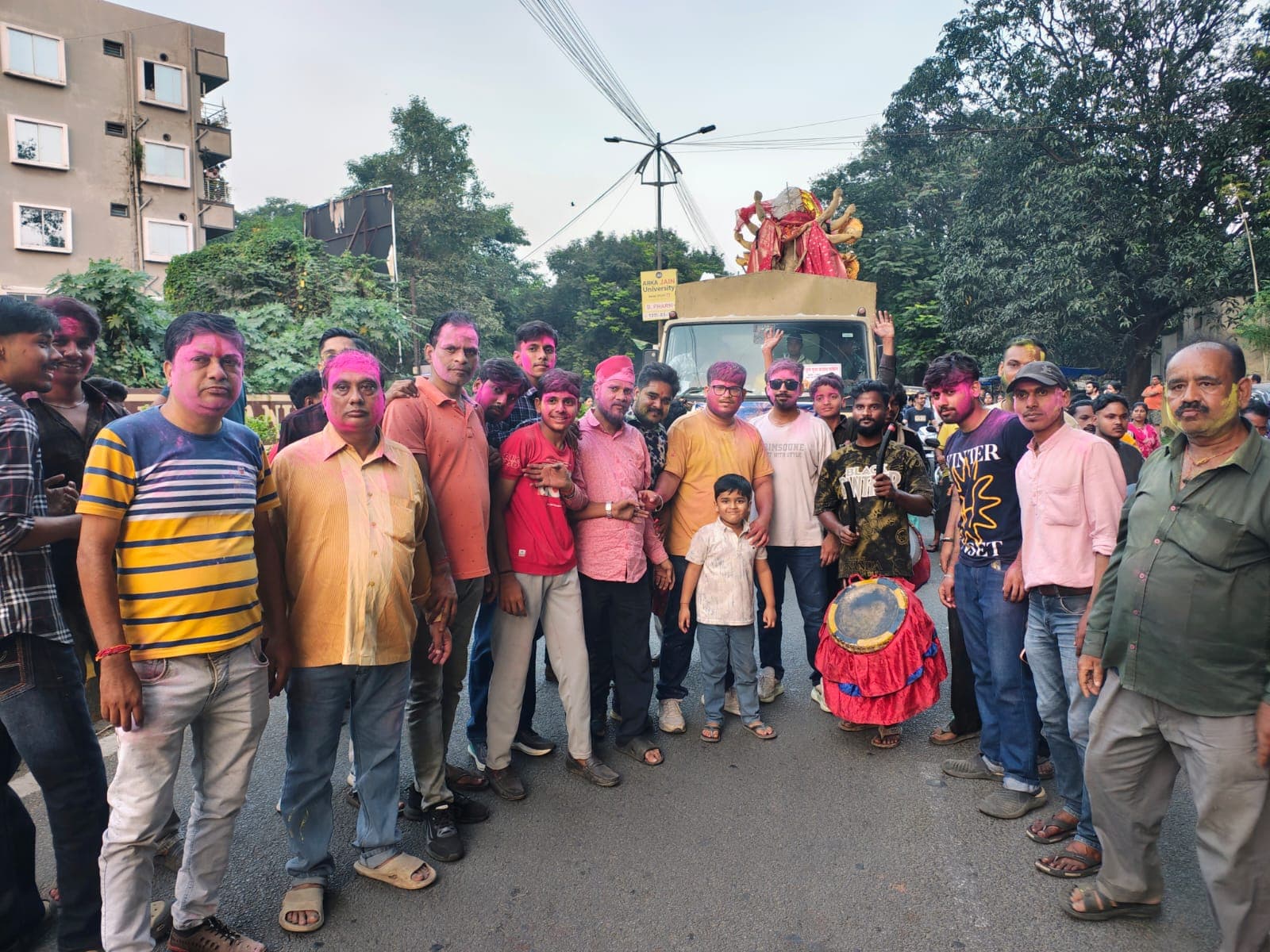 सड़क पर गुलाबी और पीले रंग से रंगे चेहरों वाले कई लोग समूह में खड़े हैं। पीछे एक ट्रक पर देवी की प्रतिमा रखी दिखाई दे रही है और एक व्यक्ति ढोल के साथ खड़ा है।