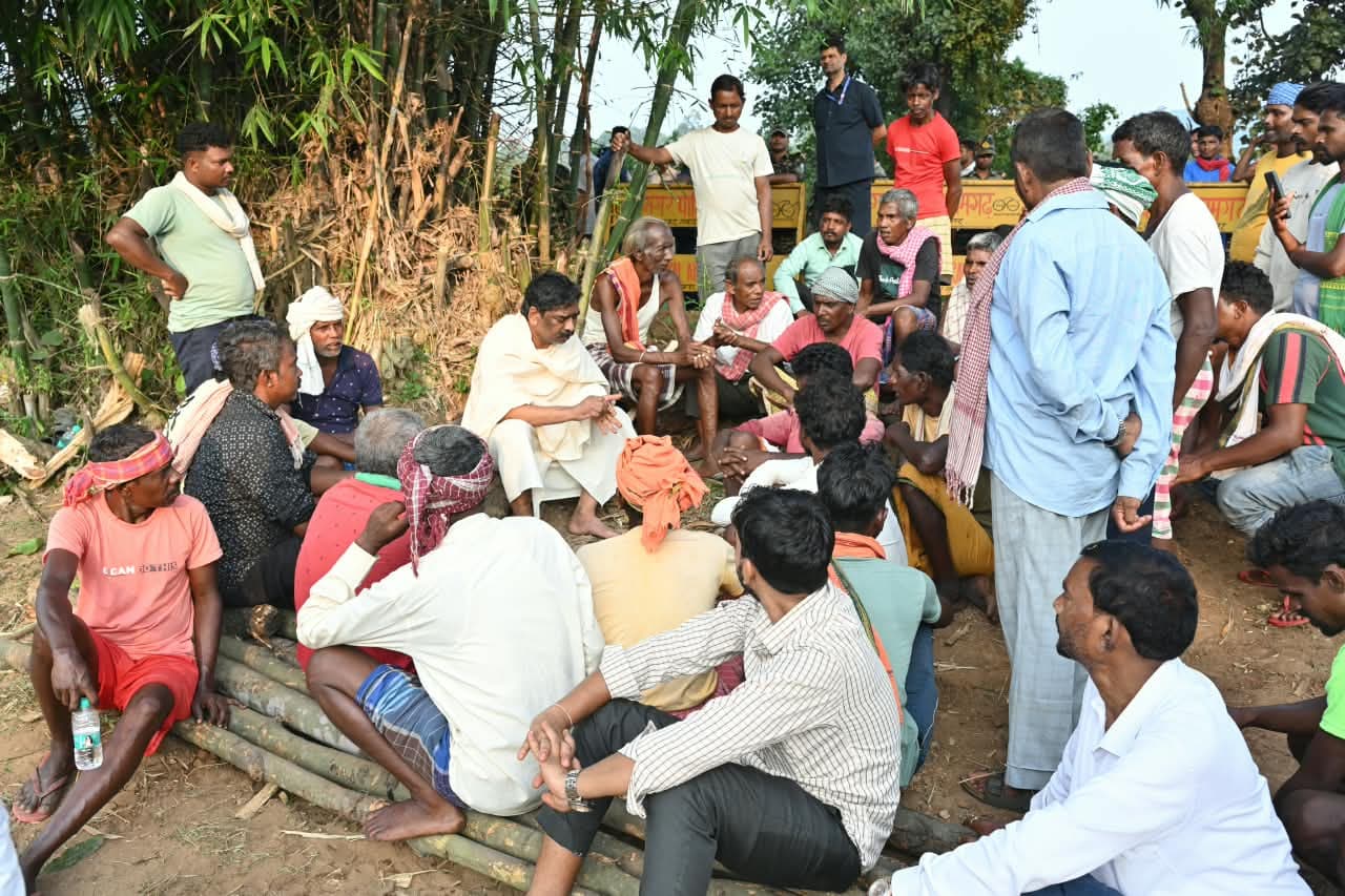 बाँस के झुरमुट के पास ज़मीन पर बैठे ग्रामीणों के बीच सफ़ेद धोती और चादर पहने एक व्यक्ति शोक की मुद्रा में बैठे हैं, चारों ओर लोग बैठे और खड़े हैं।