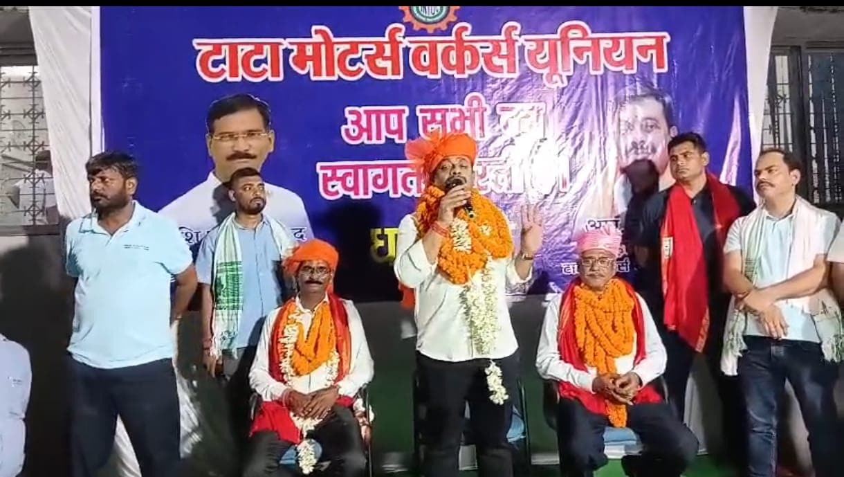 एक मंच पर कई लोग मौजूद हैं। बीच में एक व्यक्ति माइक्रोफोन के माध्यम से संबोधित कर रहा है, जबकि अन्य लोग उसके आसपास खड़े और बैठे दिखाई दे रहे हैं। पीछे लगे बड़े बैनर पर टाटा मोटर्स वर्कर्स यूनियन का नाम दिखाई दे रहा है।