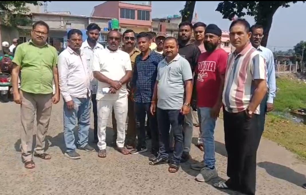 सड़क किनारे एक कतार में खड़े कई पुरुष कैमरे की ओर देख रहे हैं। उनके पीछे मकान और पेड़ दिखाई दे रहे हैं, और दाईं ओर घास तथा नाले जैसा हिस्सा है।