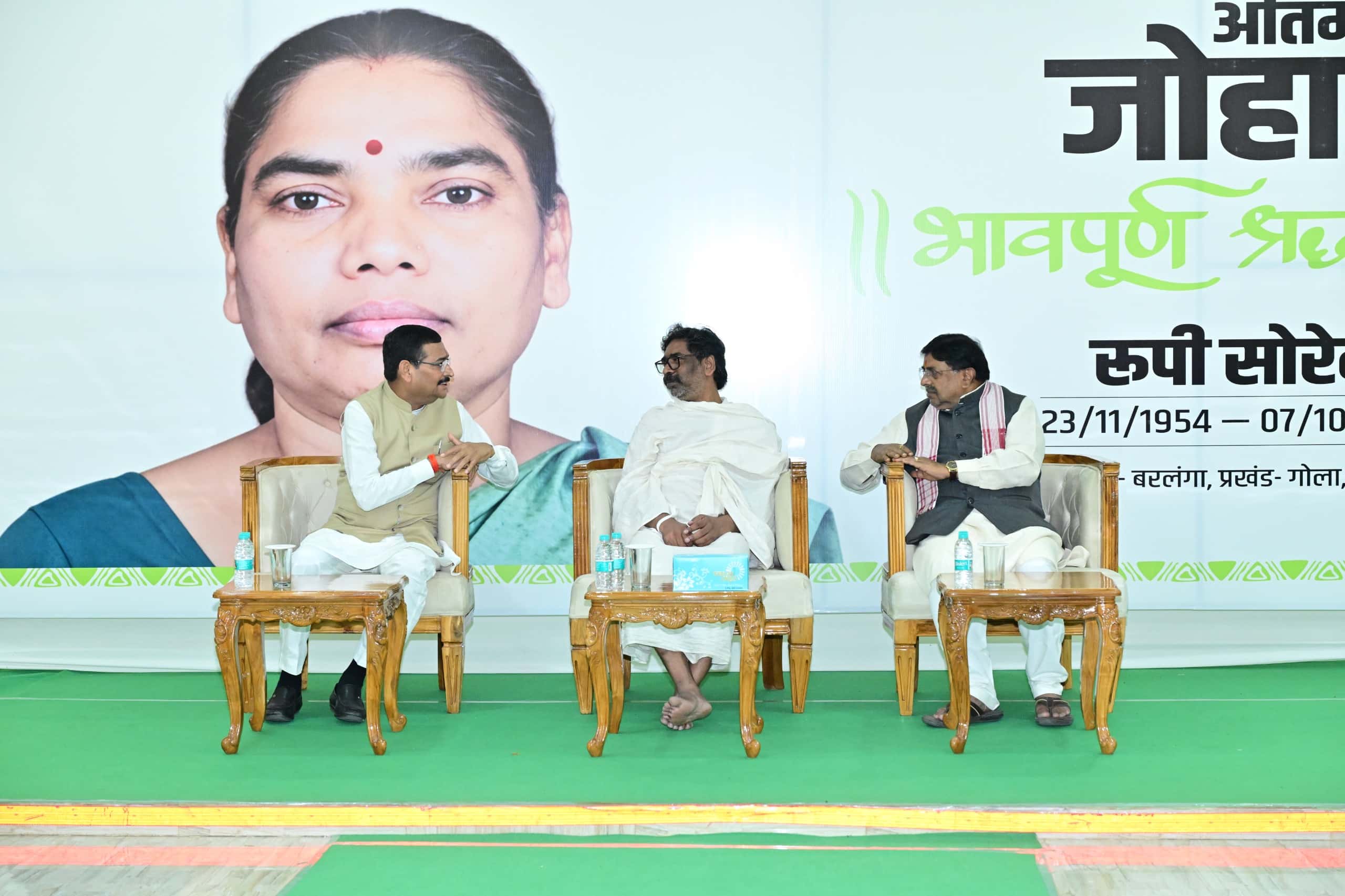 हरे कालीन वाले मंच पर तीन पुरुष गद्दीदार कुर्सियों पर बैठे हैं। बीच में सफेद चादर ओढ़े व्यक्ति के दोनों ओर बैठे लोग उनसे बातचीत करते दिख रहे हैं। पीछे बड़े बैनर पर एक महिला की तस्वीर है और हिंदी में लिखे आंशिक पाठ में रूपी सोरेन का नाम पढ़ा जा सकता है।