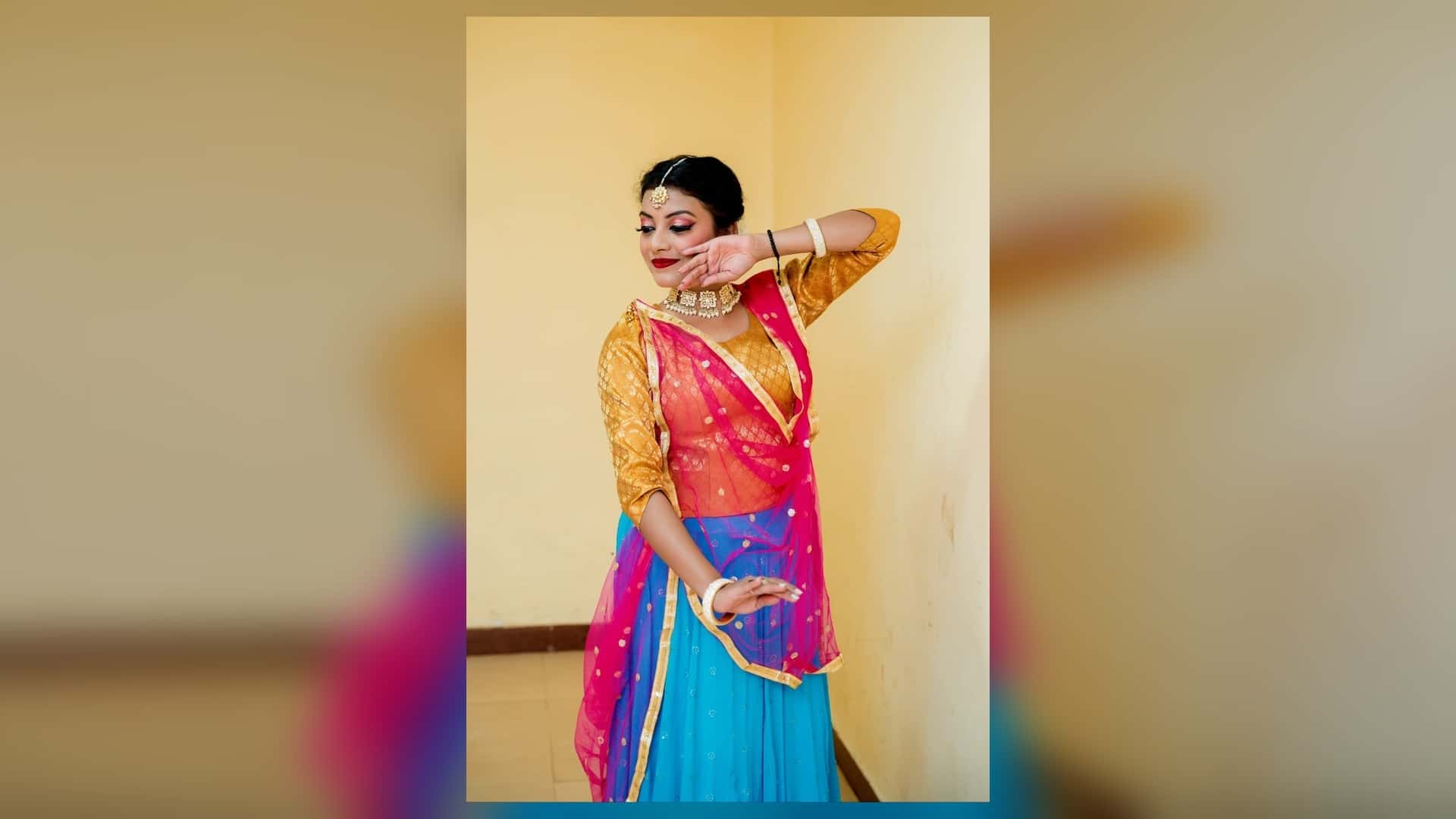 पीले-सुनहरे ब्लाउज, गुलाबी दुपट्टे और नीले लहंगे में एक महिला कथक मुद्रा में खड़ी हैं। एक हाथ चेहरे के पास और दूसरा कमर के पास है। उन्होंने मांग टीका, हार और चूड़ियां पहनी हैं और पीछे पीली दीवार दिख रही है।