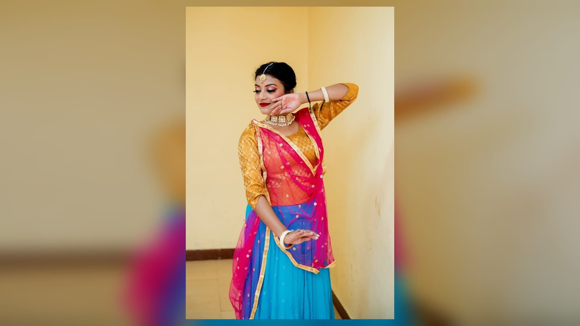 पीले-सुनहरे ब्लाउज, गुलाबी दुपट्टे और नीले लहंगे में एक महिला कथक मुद्रा में खड़ी हैं। एक हाथ चेहरे के पास और दूसरा कमर के पास है। उन्होंने मांग टीका, हार और चूड़ियां पहनी हैं और पीछे पीली दीवार दिख रही है।