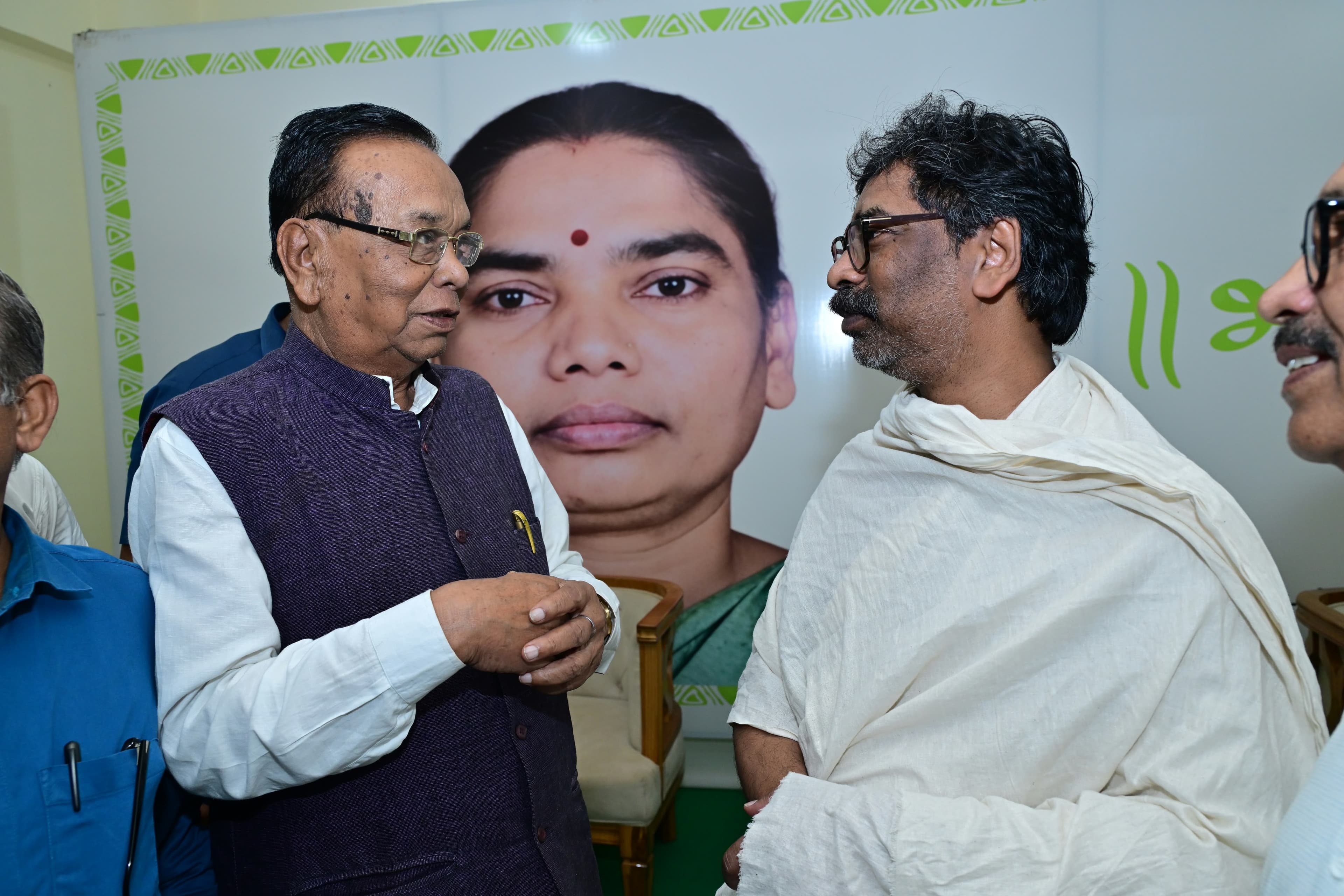 हरी जनजातीय डिज़ाइन वाली बॉर्डर के बैनर के सामने दो पुरुष आमने-सामने खड़े होकर बातचीत कर रहे हैं। एक ने बैंगनी जैकेट और सफेद कुर्ता पहना है, दूसरे ने सफेद चादर ओढ़ रखी है। पीछे बैनर पर एक महिला का बड़ा चित्र है और आसपास कुछ और लोग दिखाई दे रहे हैं।