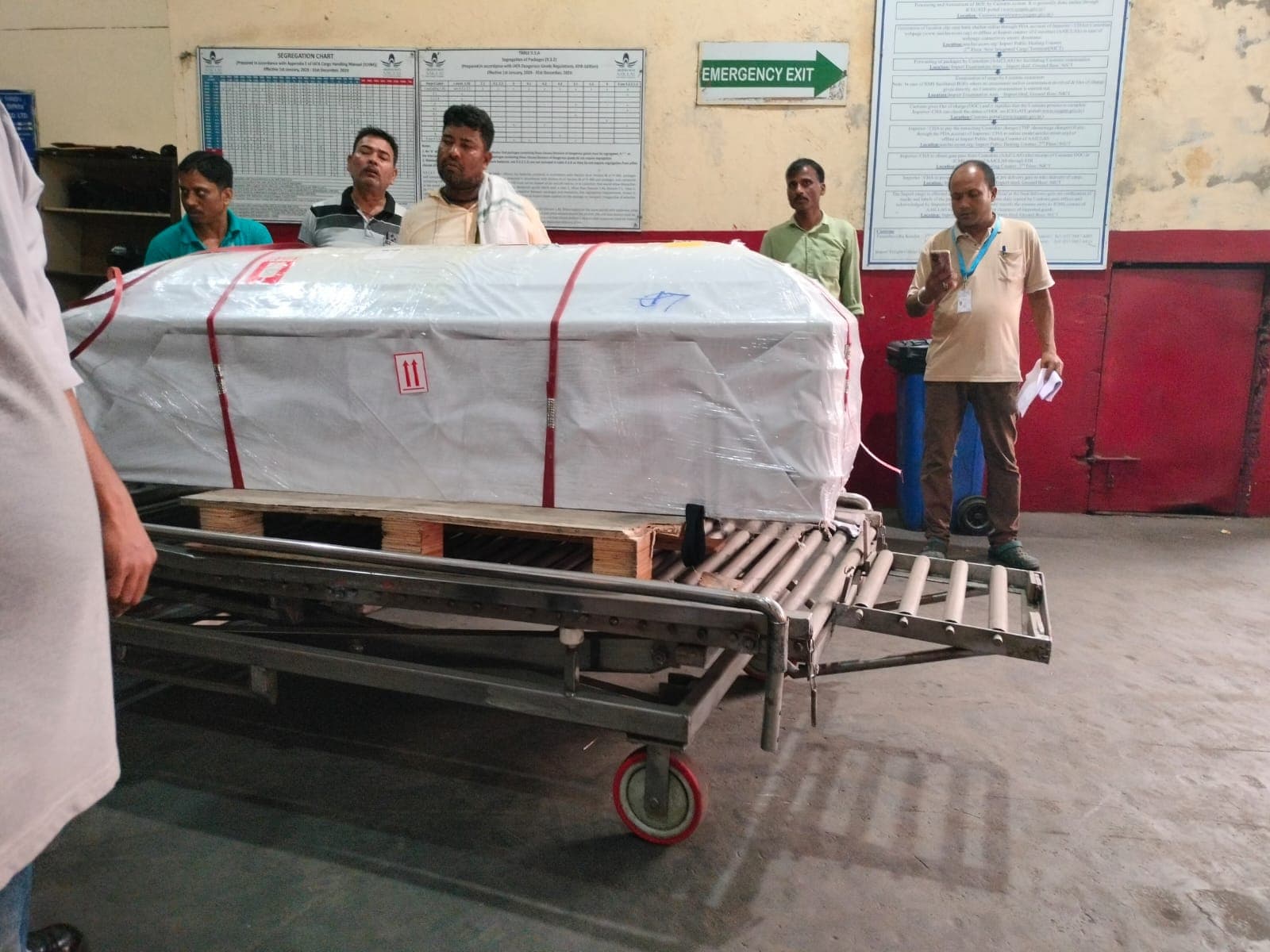 एक संस्थागत परिसर के अंदर पहियों वाली धातु की ट्रॉली पर लकड़ी के आधार के साथ रखा प्लास्टिक से ढका बड़ा पैकेज दिखाई दे रहा है। पैकेज लाल पट्टियों से बंधा है और आसपास कई लोग खड़े हैं।