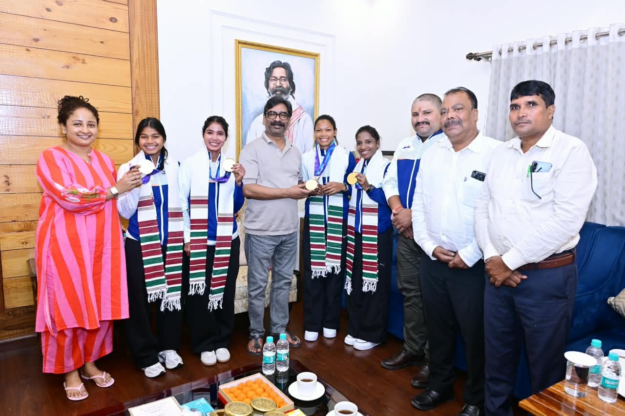 एक कमरे में कई लोग कतार में खड़े होकर तस्वीर खिंचवा रहे हैं। इनमें हरी-लाल धारियों वाला अंगवस्त्र पहने चार महिलाएं स्वर्ण पदक दिखा रही हैं। उनके साथ गुलाबी धारीदार पोशाक में एक महिला और दाईं ओर तीन पुरुष खड़े हैं।