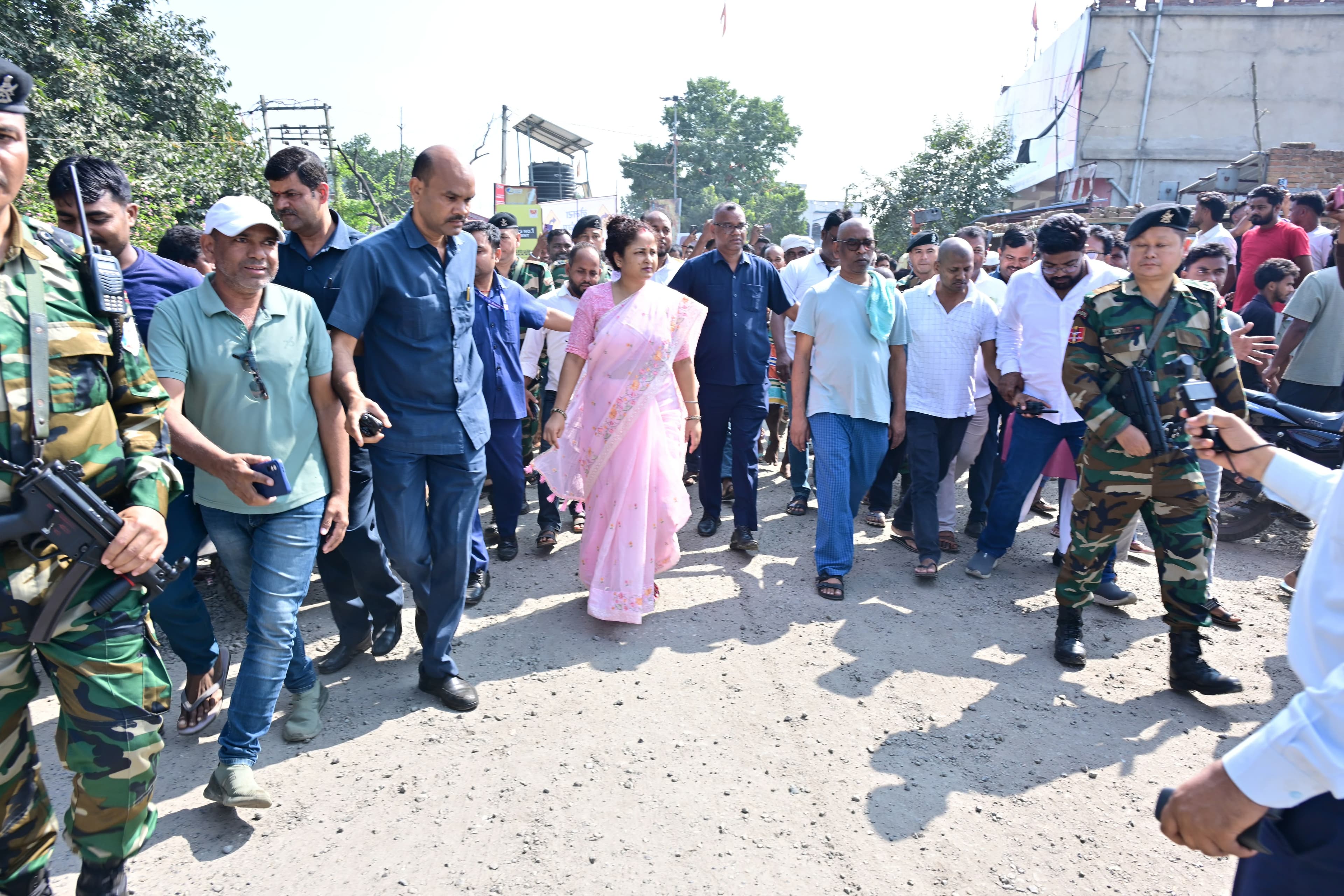 धूप में एक कच्ची-उबड़ सड़क पर गुलाबी साड़ी पहने एक महिला के साथ चलती भीड़ दिखाई दे रही है। आसपास कैमोफ्लाज वर्दी में हथियारबंद सुरक्षाकर्मी और कई स्थानीय लोग हैं।