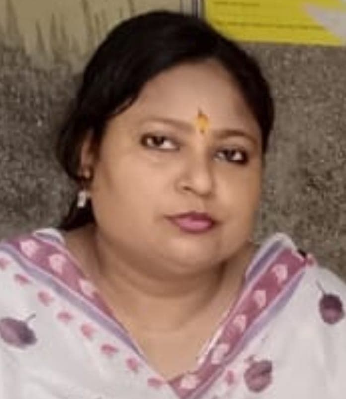 एक महिला का क्लोज-अप चित्र। उसके काले बाल पीछे बंधे हैं, माथे पर पीली-नारंगी बिंदी है और कान में सफेद बाली है। उसने गुलाबी-बैंगनी किनारी और छपाई वाला सफेद परिधान पहना है। पीछे पत्थर जैसी दीवार दिखती है।