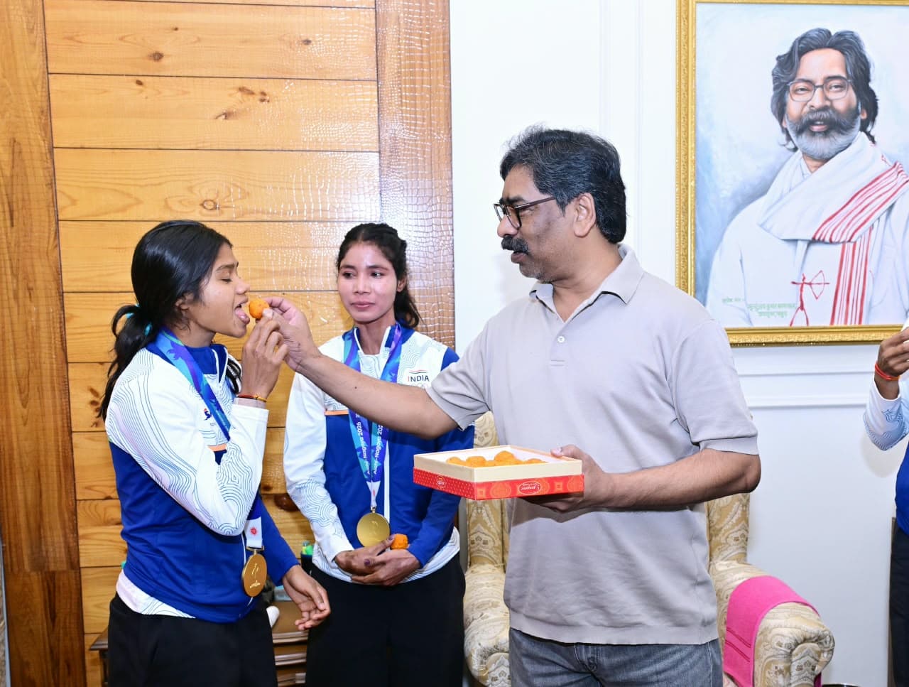 एक पुरुष मिठाई के डिब्बे से मिठाई उठाकर गले में पदक पहने एक महिला को खिला रहे हैं। आसपास नीली जैकेट पहने कुछ और लोग मुस्कुराते हुए खड़े हैं।