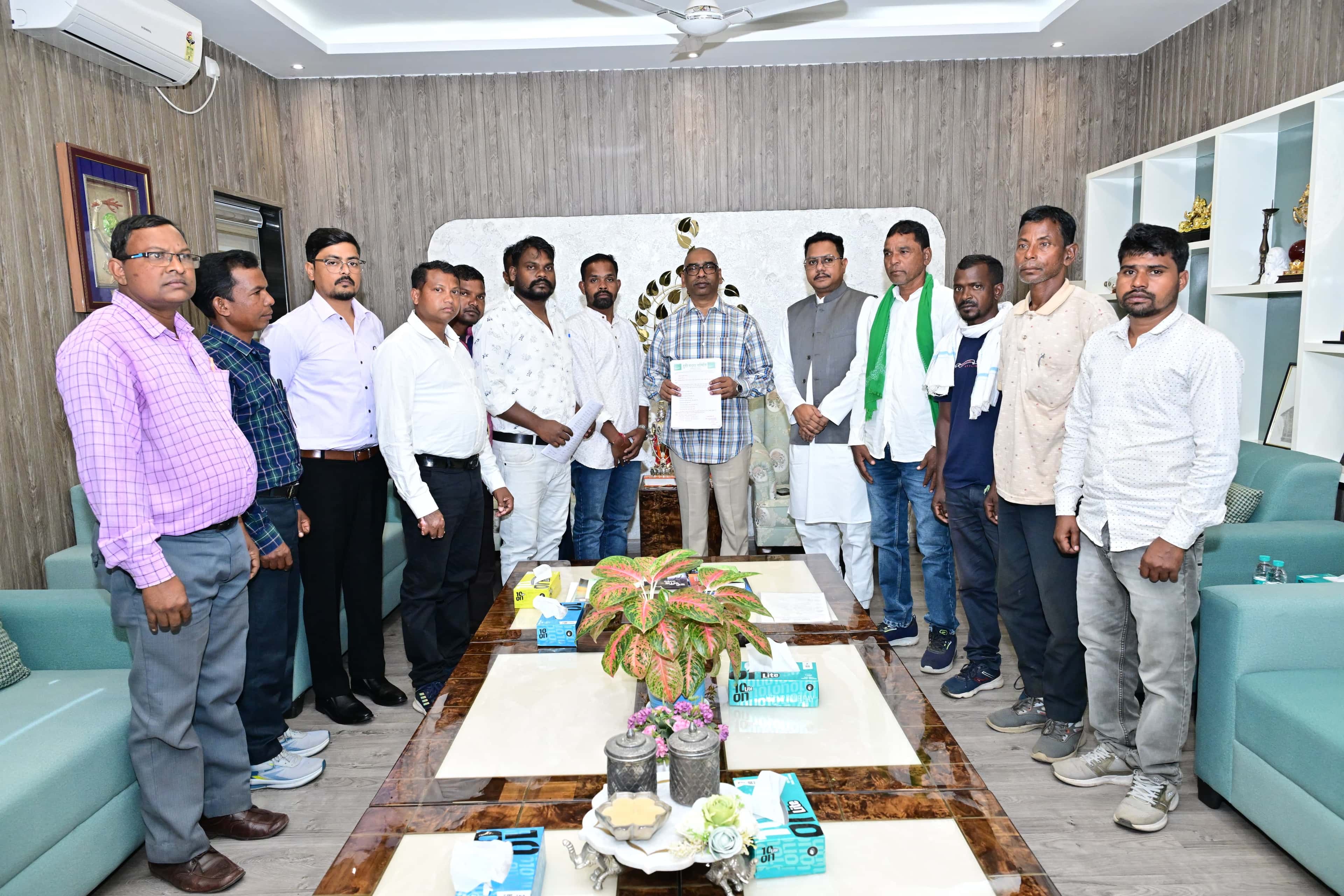 लकड़ी जैसी दीवार पैनलिंग वाले कमरे में 12-13 पुरुष एक पंक्ति में खड़े हैं। बीच में खड़े एक व्यक्ति के हाथ में हरे शीर्षक वाला कागज है। एक व्यक्ति के गले में हरा गमछा है। आगे मेज पर गमले में पौधा और टिश्यू बॉक्स रखे हैं।