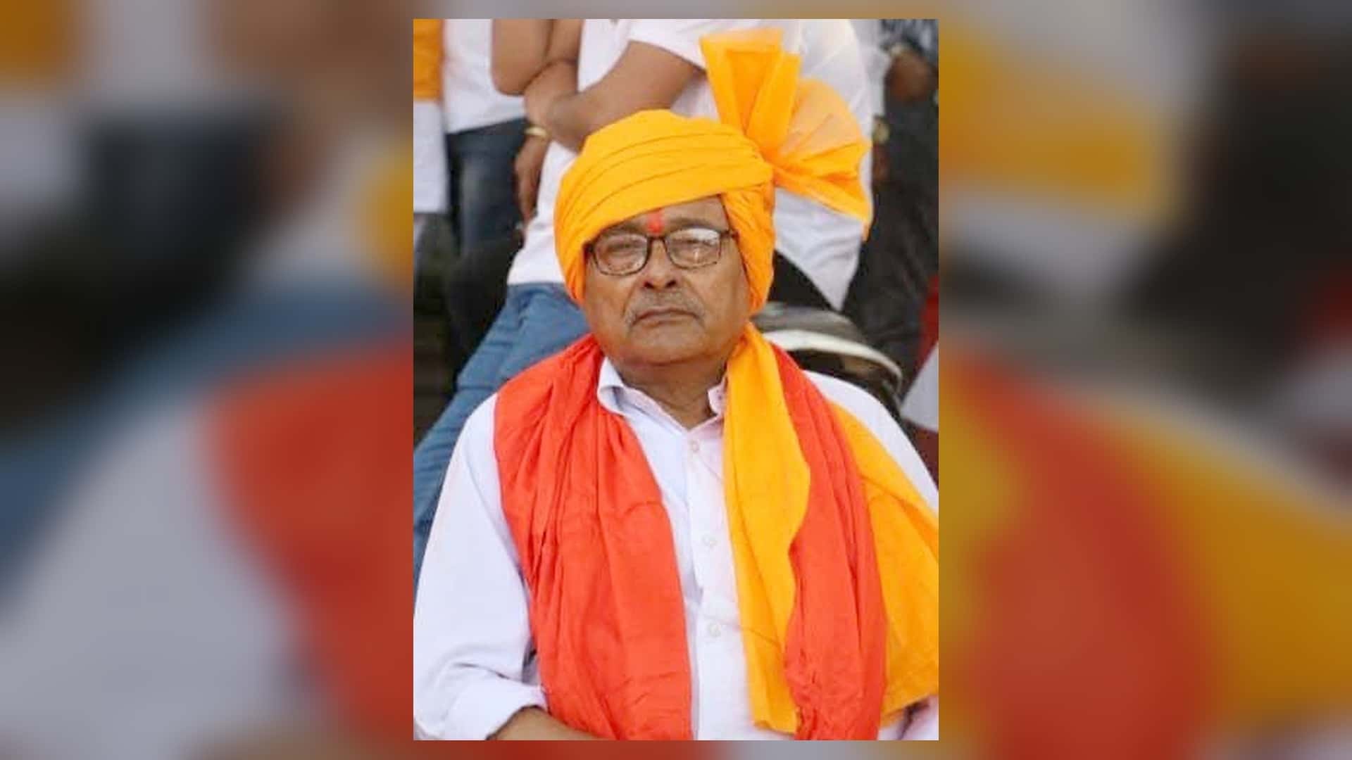 केसरिया-पीले रंग की पगड़ी और नारंगी गमछा पहने, चश्मा लगाए एक बुजुर्ग व्यक्ति के माथे पर तिलक दिखाई दे रहा है। उन्होंने सफेद शर्ट पहनी है और पीछे कुछ लोग बैठे नजर आ रहे हैं।