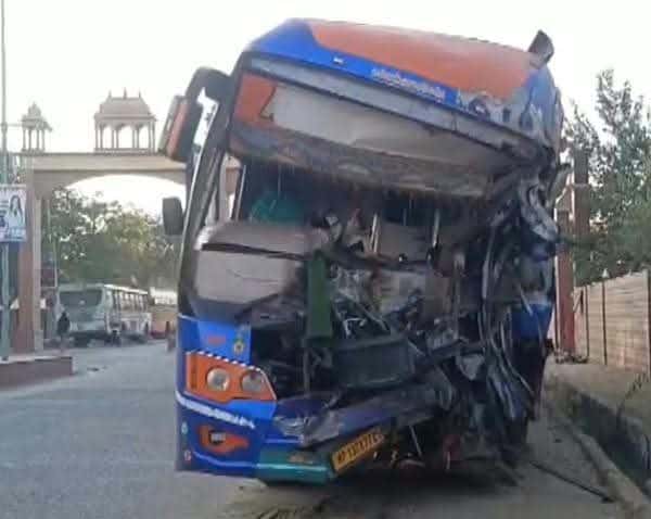 सड़क किनारे खड़ी एक नीली-नारंगी बस का अगला हिस्सा बुरी तरह टूटा-पिचका दिख रहा है। शीशे और ढांचा क्षतिग्रस्त है। पीछे एक द्वार जैसी संरचना और अन्य वाहन दिखाई दे रहे हैं।