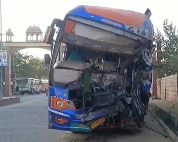 सड़क किनारे खड़ी एक नीली-नारंगी बस का अगला हिस्सा बुरी तरह टूटा-पिचका दिख रहा है। शीशे और ढांचा क्षतिग्रस्त है। पीछे एक द्वार जैसी संरचना और अन्य वाहन दिखाई दे रहे हैं।