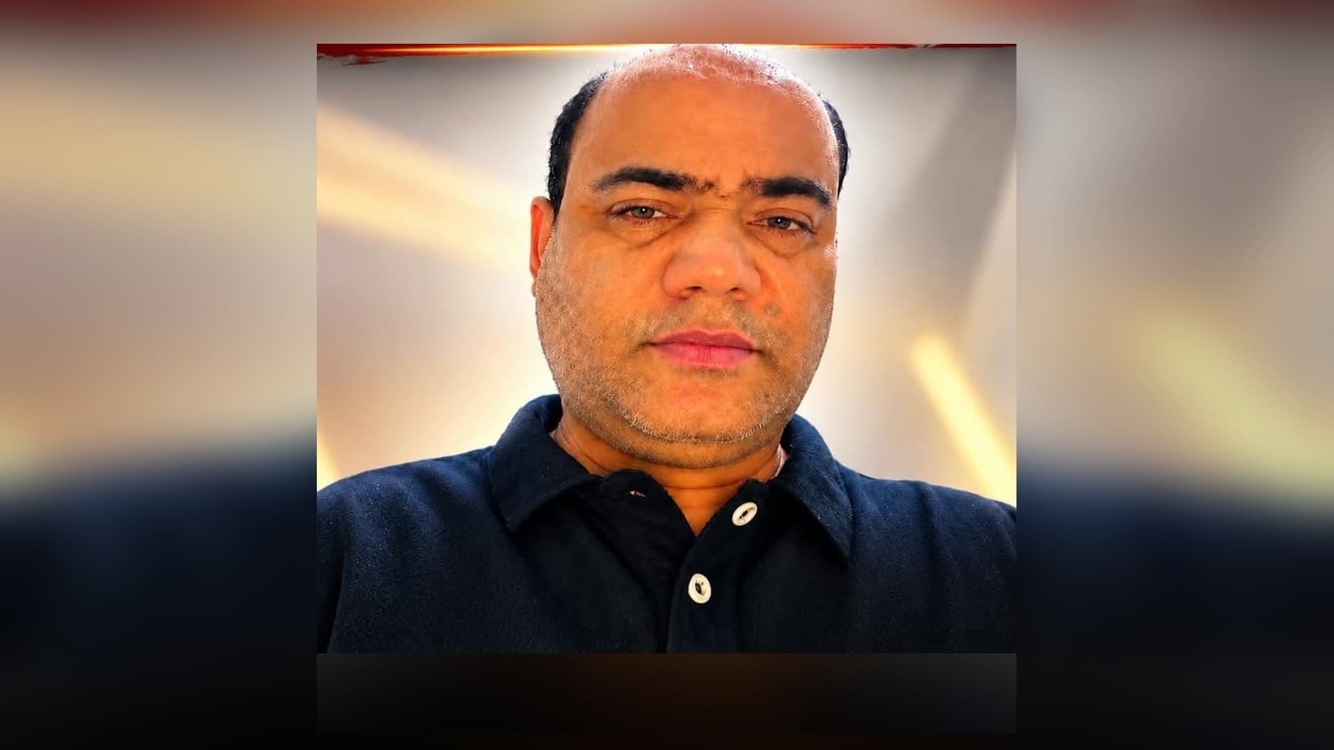 गहरे नीले रंग की कॉलर वाली टी-शर्ट पहने, हल्की दाढ़ी वाला एक व्यक्ति कैमरे की ओर देख रहा है। पीछे हल्की पृष्ठभूमि पर सुनहरी रोशनी की धारियां दिखाई दे रही हैं।