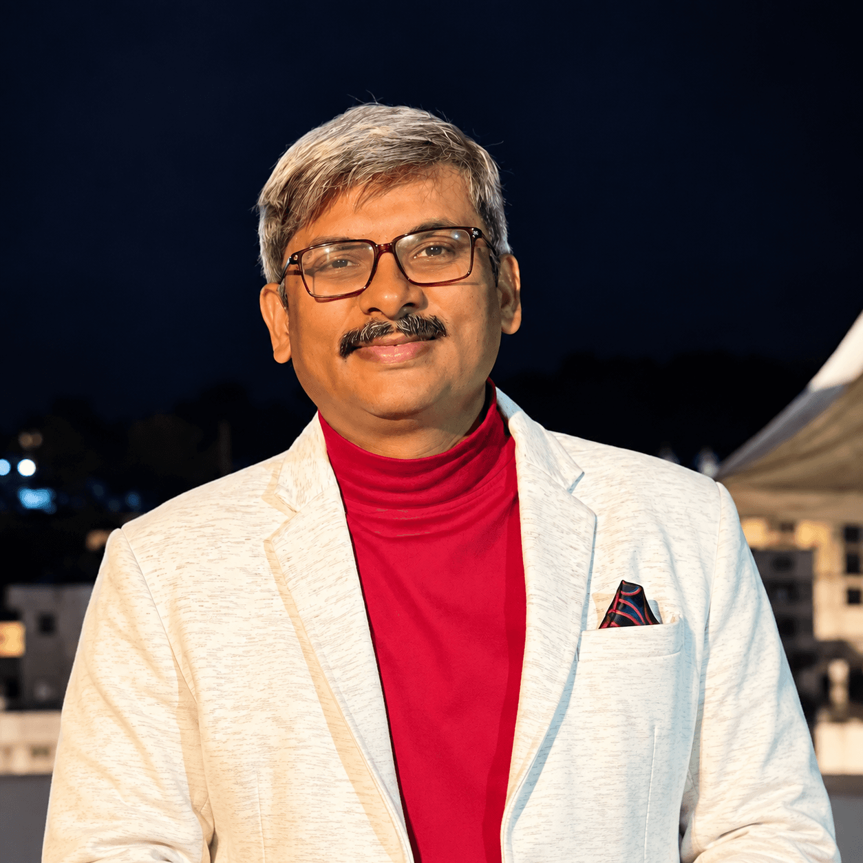 एक व्यक्ति सफेद जैकेट और लाल रंग की शर्ट पहने कैमरे की ओर देखते हुए दिखाई दे रहा है। तस्वीर एक इनडोर कार्यक्रम या मंच के दौरान ली गई प्रतीत होती है और व्यक्ति के पीछे गहरे रंग की पृष्ठभूमि दिखाई दे रही है।