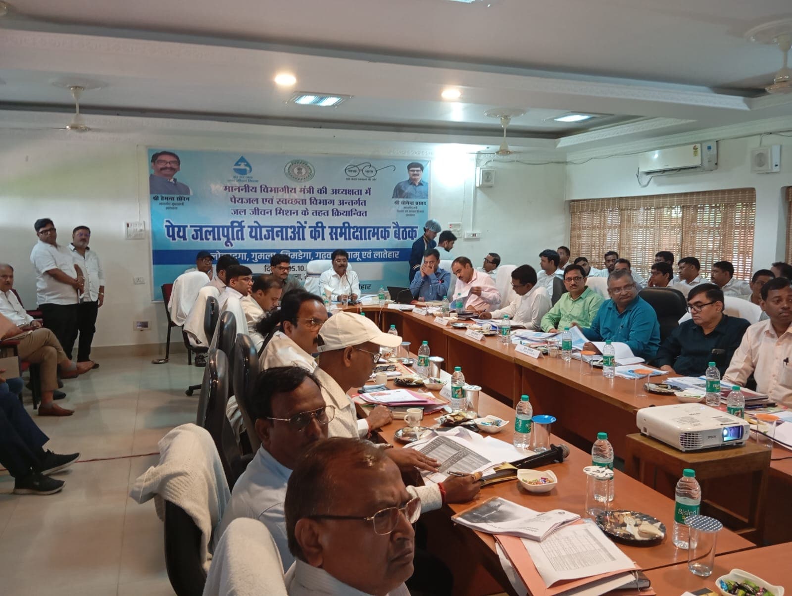 बैठक कक्ष में मेज के दोनों ओर कागजात और फाइलों के साथ बैठे कई अधिकारी। मेज पर प्रोजेक्टर रखा है और पीछे जल जीवन मिशन का बैनर लगा है।