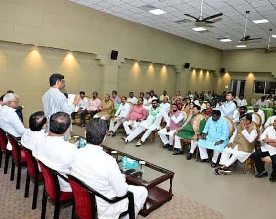 एक बड़े सभागार में कुर्सियों पर बैठे कई लोग एक खड़े व्यक्ति को सुन रहे हैं। सामने कांच की मेज और लाल कुर्सियां हैं, छत पर पंखे और लाइटें लगी हैं।