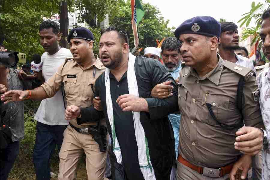 काला कुर्ता और सफेद-हरे रंग का गमछा पहने एक व्यक्ति तेजी से चलते हुए दिख रहे हैं। उनके दोनों ओर खाकी वर्दी और नीली टोपी पहने पुलिसकर्मी हैं और पीछे लोगों की भीड़ तथा एक झंडा नजर आता है। बाईं ओर कैमरा और माइक्रोफोन लिए मीडियाकर्मी मौजूद हैं।