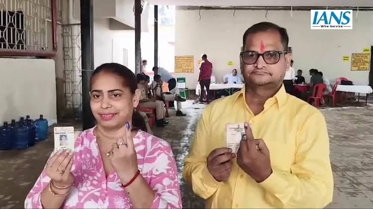 एक महिला और पुरुष हाथों में कार्ड और स्याही लगी उंगली दिखाते हुए। पीछे हॉल में मेजों के पास कई लोग बैठे हैं।