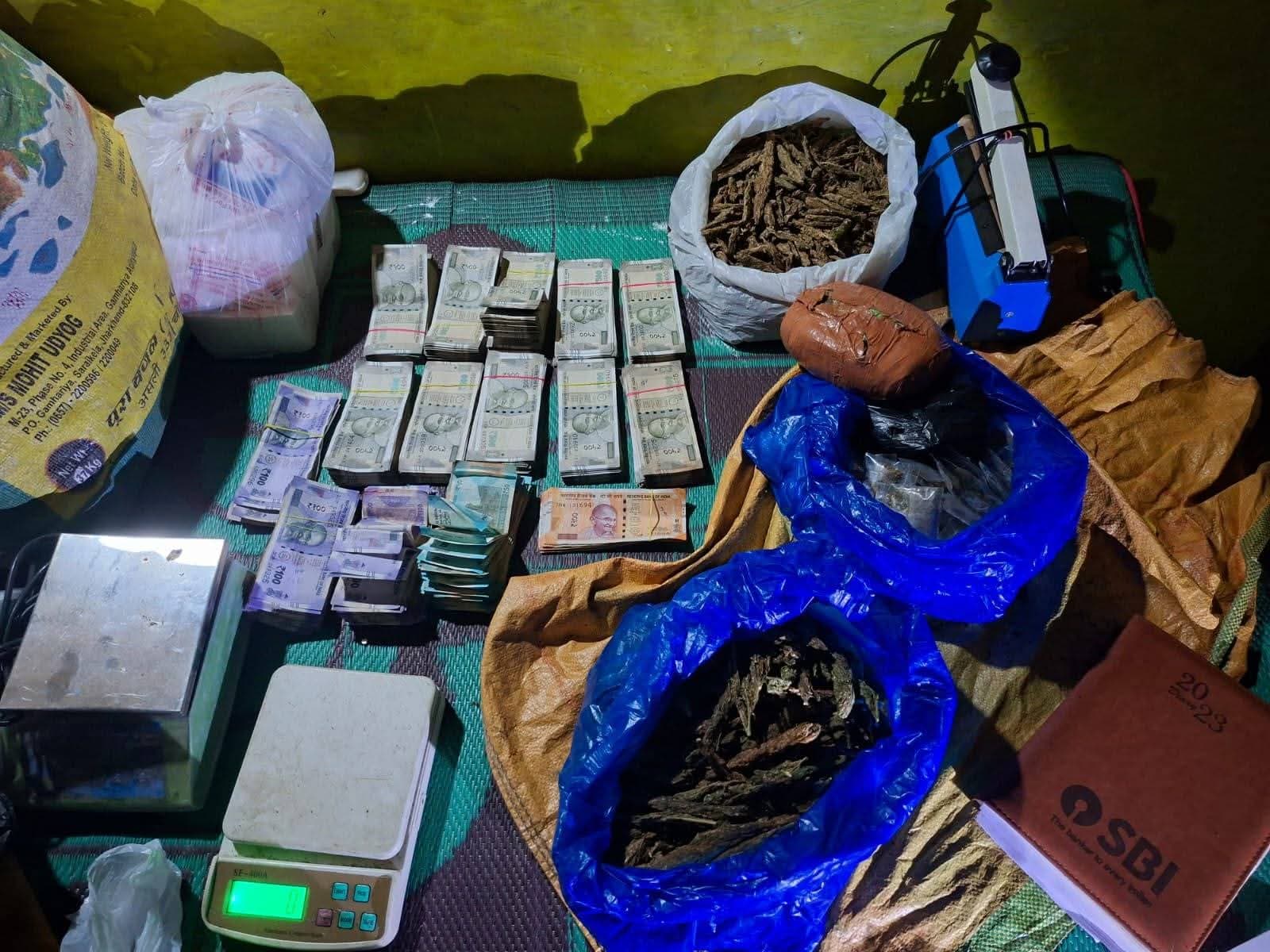 एक कमरे में रखी चारपाई पर नकदी के कई बंडल, weighing scales और पॉलीथिन में रखा सूखा पदार्थ दिखाई दे रहा है। तस्वीर में बरामद सामान को एक साथ रखा गया है।