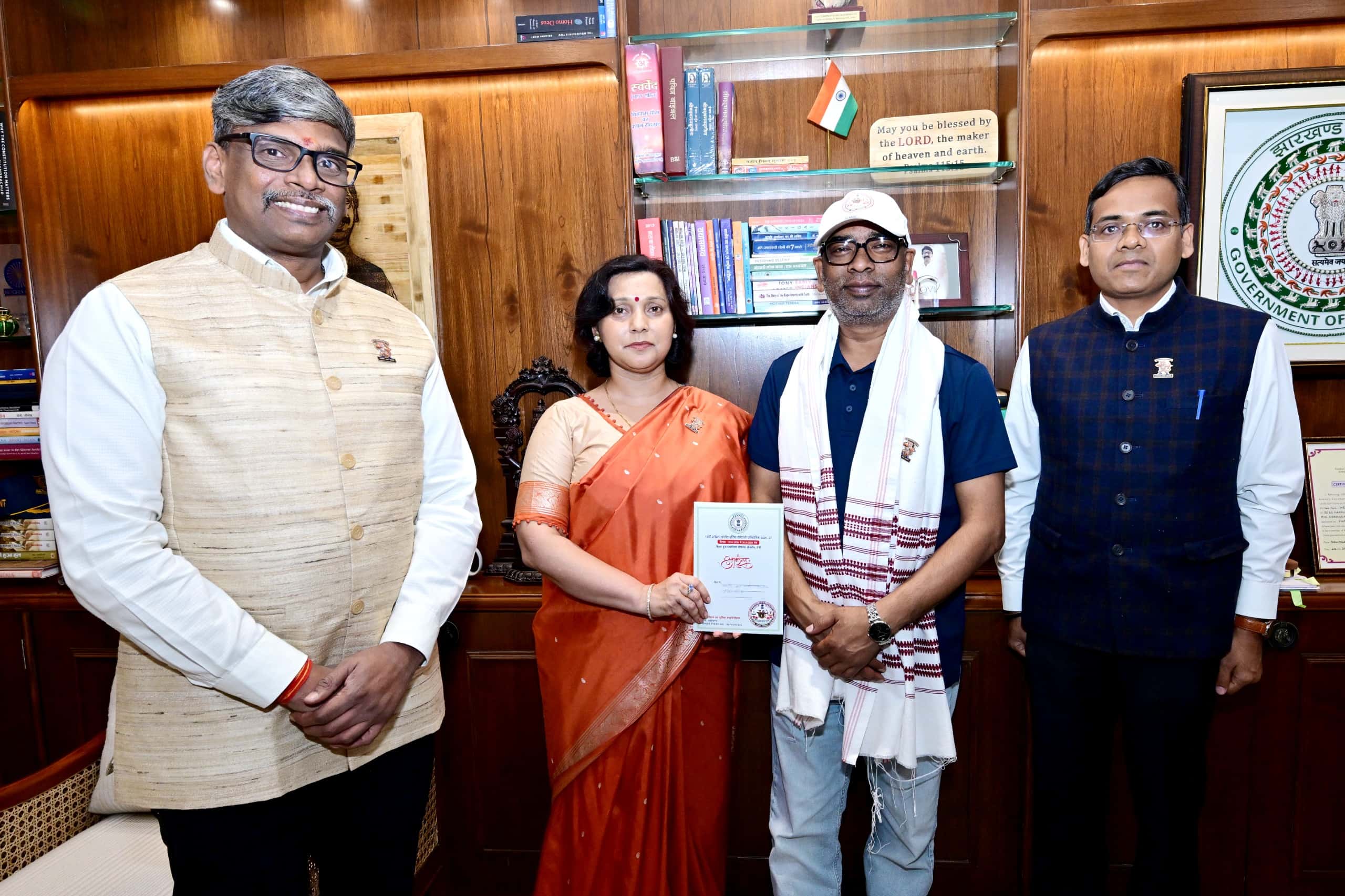 लकड़ी की पैनलिंग वाले कार्यालय कक्ष में चार लोग कैमरे की ओर देखते हुए खड़े हैं। नारंगी साड़ी पहनी एक महिला हाथ में आमंत्रण पत्र जैसा कार्ड थामे हैं। पीछे अलमारी में किताबें, तिरंगा और झारखंड सरकार का प्रतीक चिह्न दिखाई दे रहे हैं।