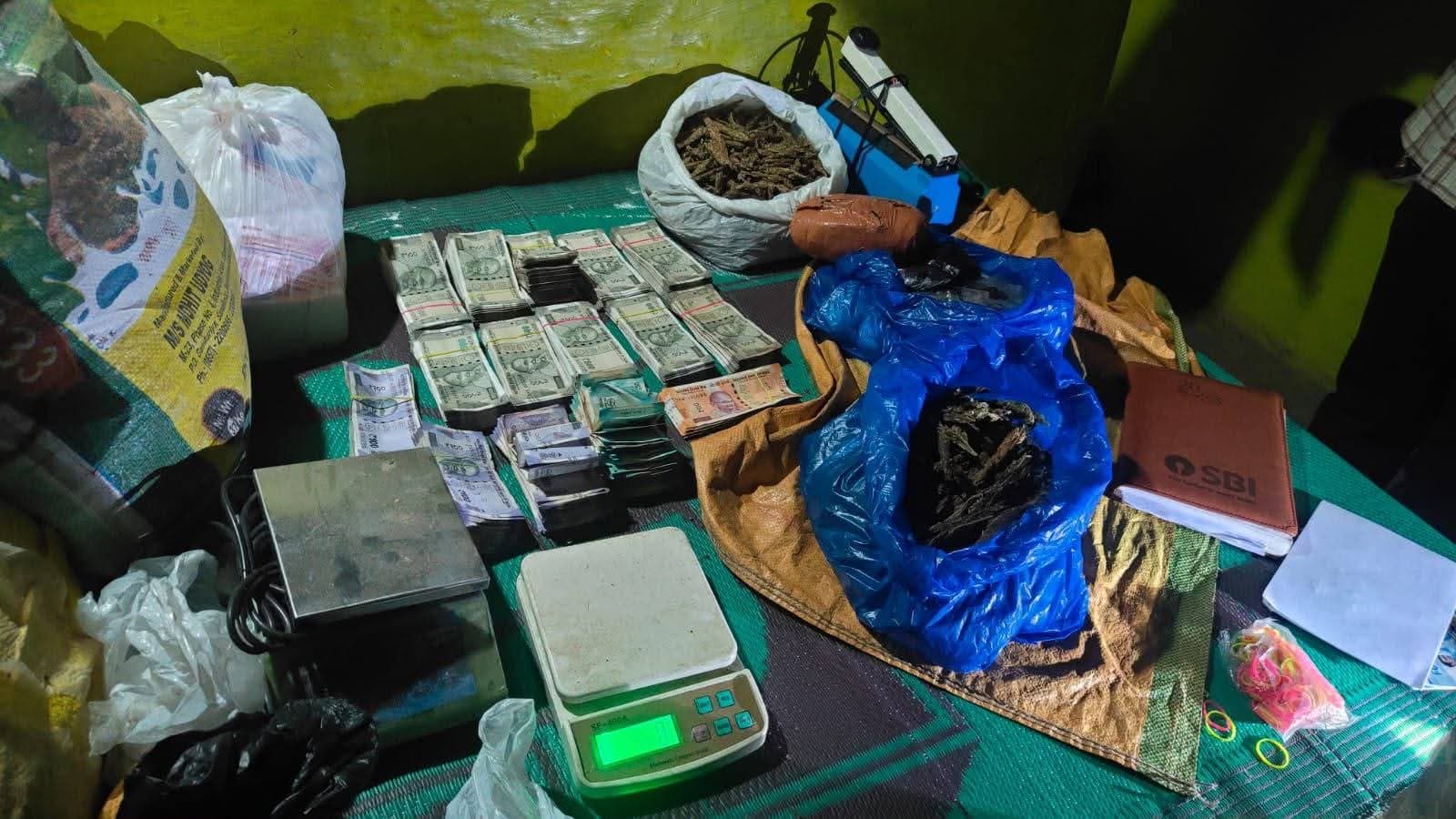 एक कमरे में रखी चारपाई पर बड़ी मात्रा में नकदी के बंडल, दो इलेक्ट्रॉनिक weighing scales और पॉलीथिन में रखा सूखा पदार्थ दिखाई दे रहा है। आसपास कुछ पैकेट और अन्य सामान भी रखा है।