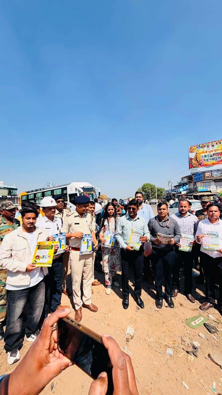 धूल भरी खुली सड़क पर खड़े कई पुरुष और एक महिला हाथों में रंगीन पंपलेट लिए दिख रहे हैं। कुछ लोग खाकी वर्दी में हैं, एक व्यक्ति ने सफेद हेलमेट पहना है। पीछे एक बस और सड़क किनारे दुकानें हैं।