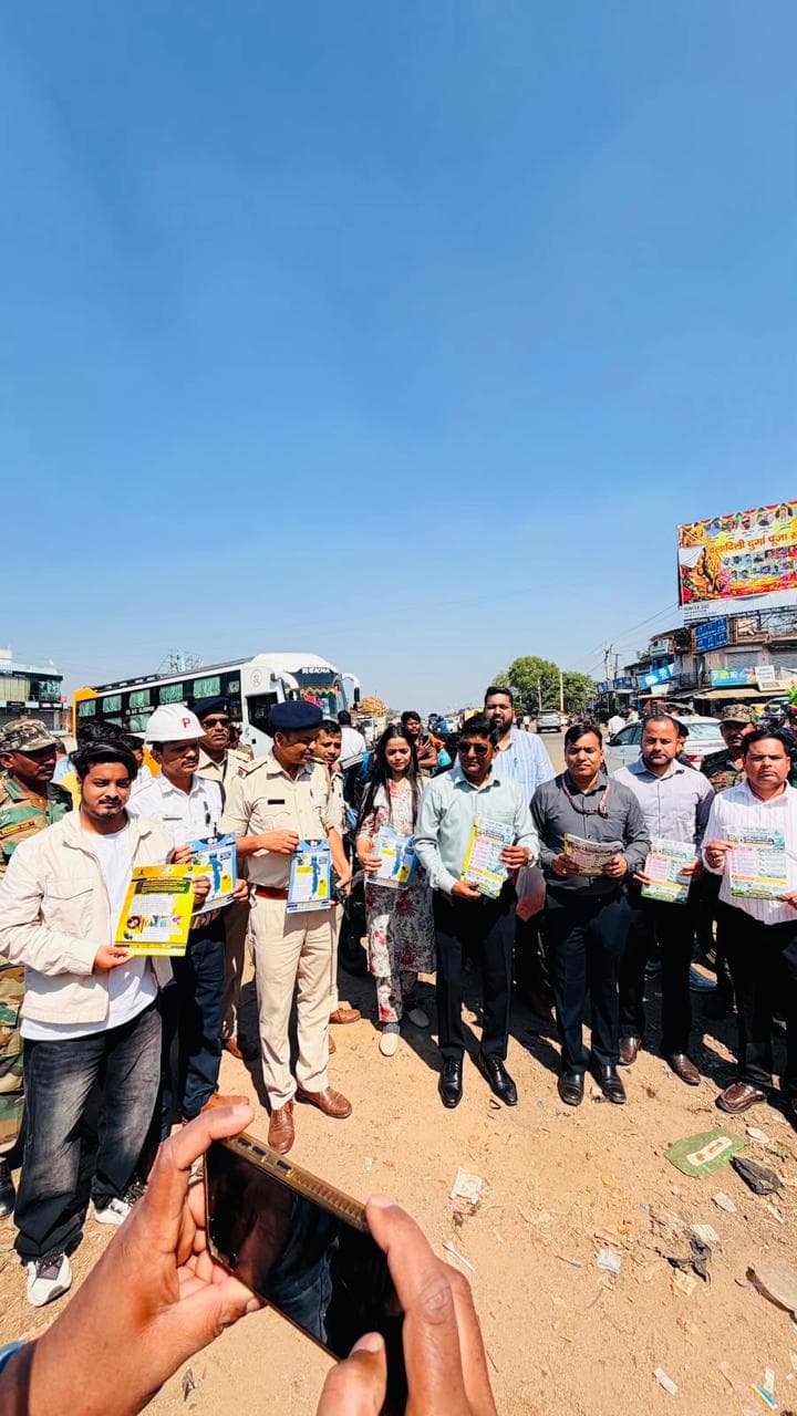 धूल भरी खुली सड़क पर खड़े कई पुरुष और एक महिला हाथों में रंगीन पंपलेट लिए दिख रहे हैं। कुछ लोग खाकी वर्दी में हैं, एक व्यक्ति ने सफेद हेलमेट पहना है। पीछे एक बस और सड़क किनारे दुकानें हैं।