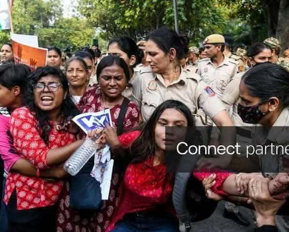 सड़क पर महिला पुलिसकर्मी कई प्रदर्शनकारी महिलाओं को पकड़े हुए दिख रही हैं। एक महिला के हाथ में पानी की बोतल और कागज हैं और वह चिल्लाती दिख रही है। पीछे पेड़ और कई लोग हैं।