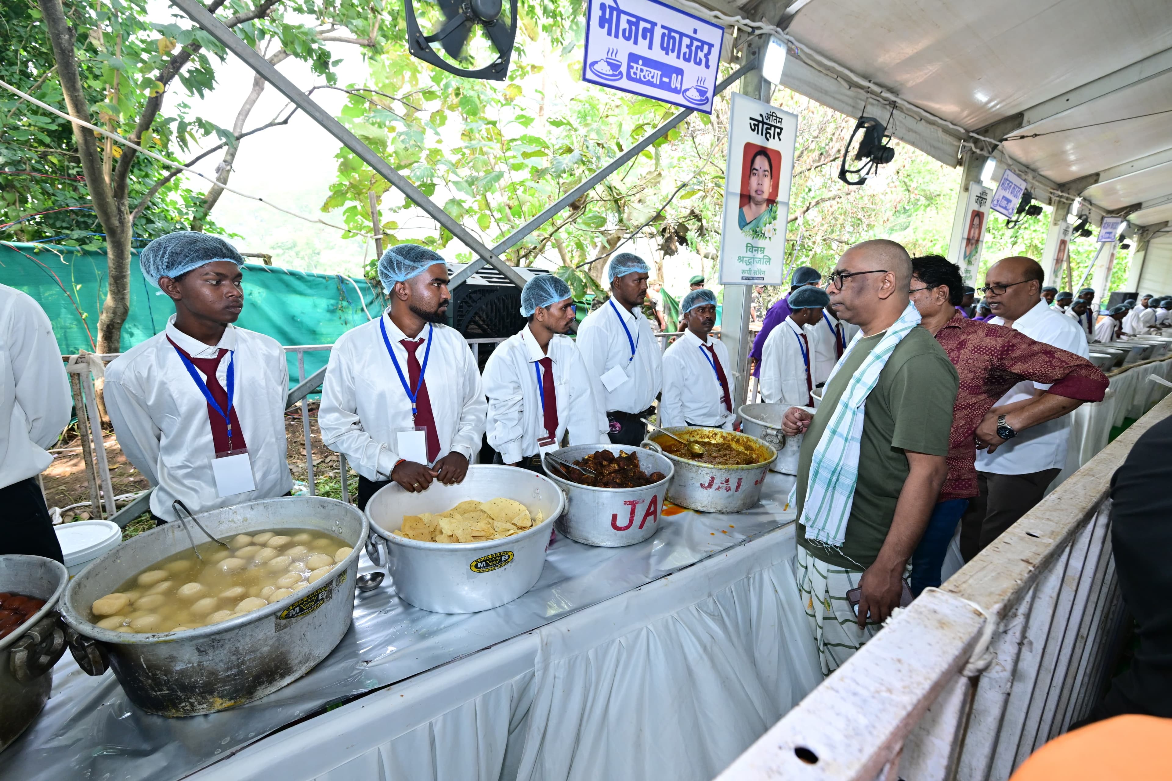 सफेद शर्ट, लाल टाई और हेयर कैप पहने कई कर्मी भोजन काउंटर पर रसगुल्ले, पापड़ और अन्य व्यंजनों के बड़े बर्तनों के पीछे खड़े हैं। सामने हरी टी-शर्ट और चेक वाला गमछा पहने एक व्यक्ति उनसे बात कर रहे हैं, पास में अन्य लोग खड़े हैं। ऊपर "भोजन काउंटर संख्या-04" का बोर्ड और श्रद्धांजलि पोस्टर लगा है।