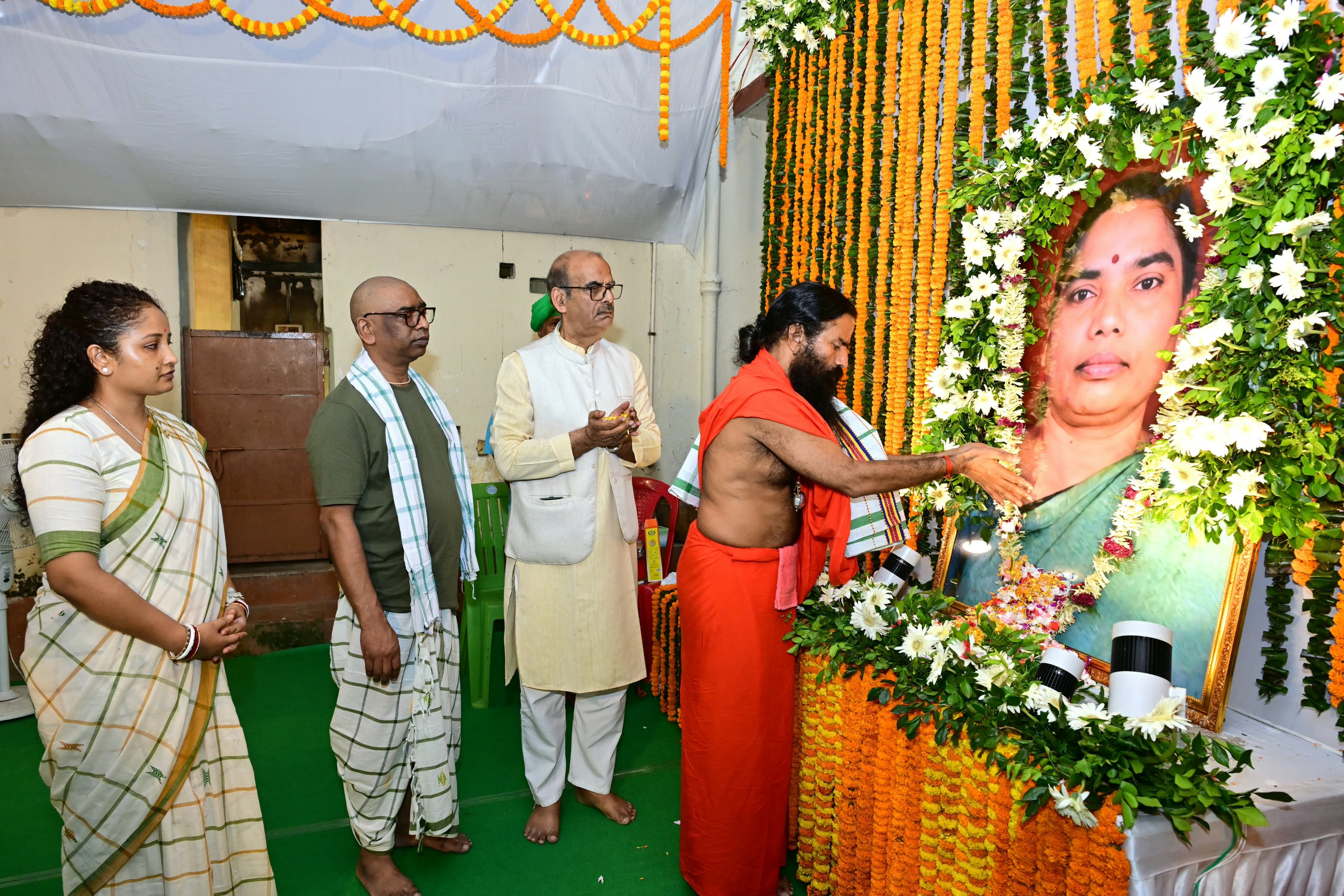 फूलों और गेंदे की मालाओं से सजे मंच पर एक महिला की फ्रेम की हुई तस्वीर पर भगवा वस्त्र पहने दाढ़ी वाला व्यक्ति पुष्प अर्पित करता दिखाई दे रहा है। पास में तीन लोग हाथ जोड़े और हाथ में फूल लिए खड़े हैं।