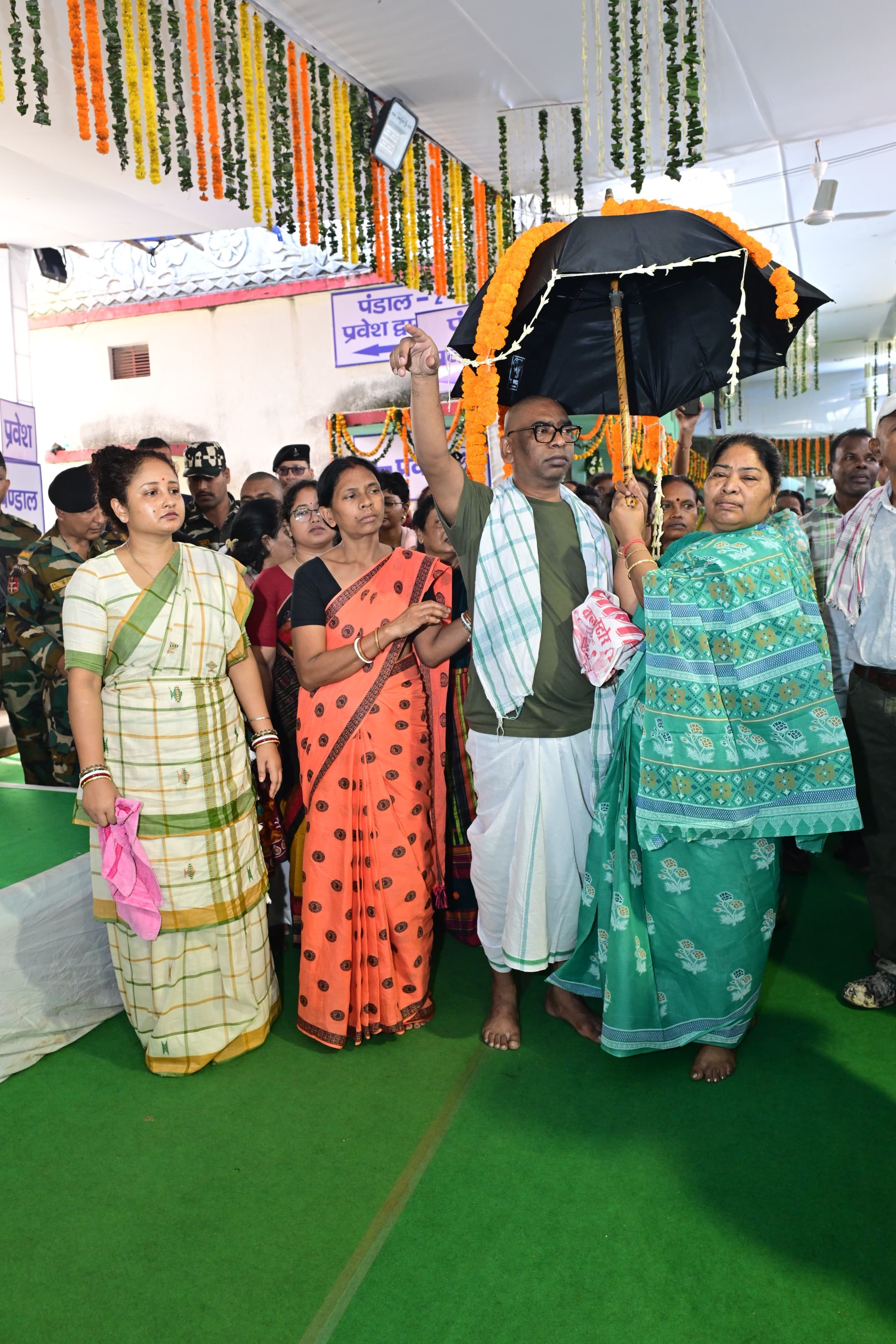 फूलों की मालाओं से सजे पंडाल में हरे कार्पेट पर मुंडित सिर और चश्मे वाला एक व्यक्ति हरी टी-शर्ट और सफेद धोती में हाथ उठाकर इशारा करता दिख रहा है। हरी साड़ी पहने एक महिला उसके ऊपर गेंदे के फूलों से सजा काला छाता थामे है। बगल में दो महिलाएं खड़ी हैं और पीछे वर्दीधारी जवानों समेत कई लोग मौजूद हैं।