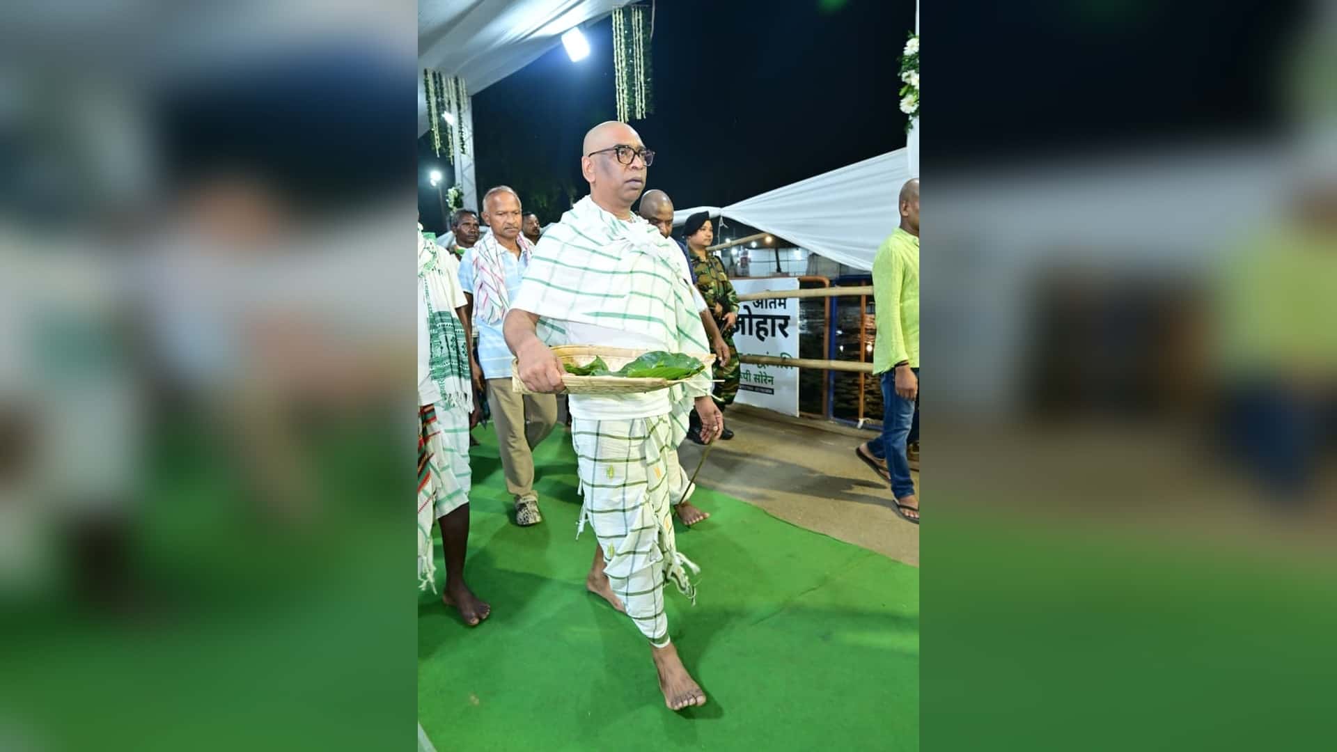 रात में हरे कालीन पर सफेद-हरी चेक वाले वस्त्र पहने एक चश्माधारी व्यक्ति नंगे पैर बांस की थाली में हरे पत्ते लेकर चलते हुए। पीछे कई लोग और सुरक्षाकर्मी दिखाई दे रहे हैं।