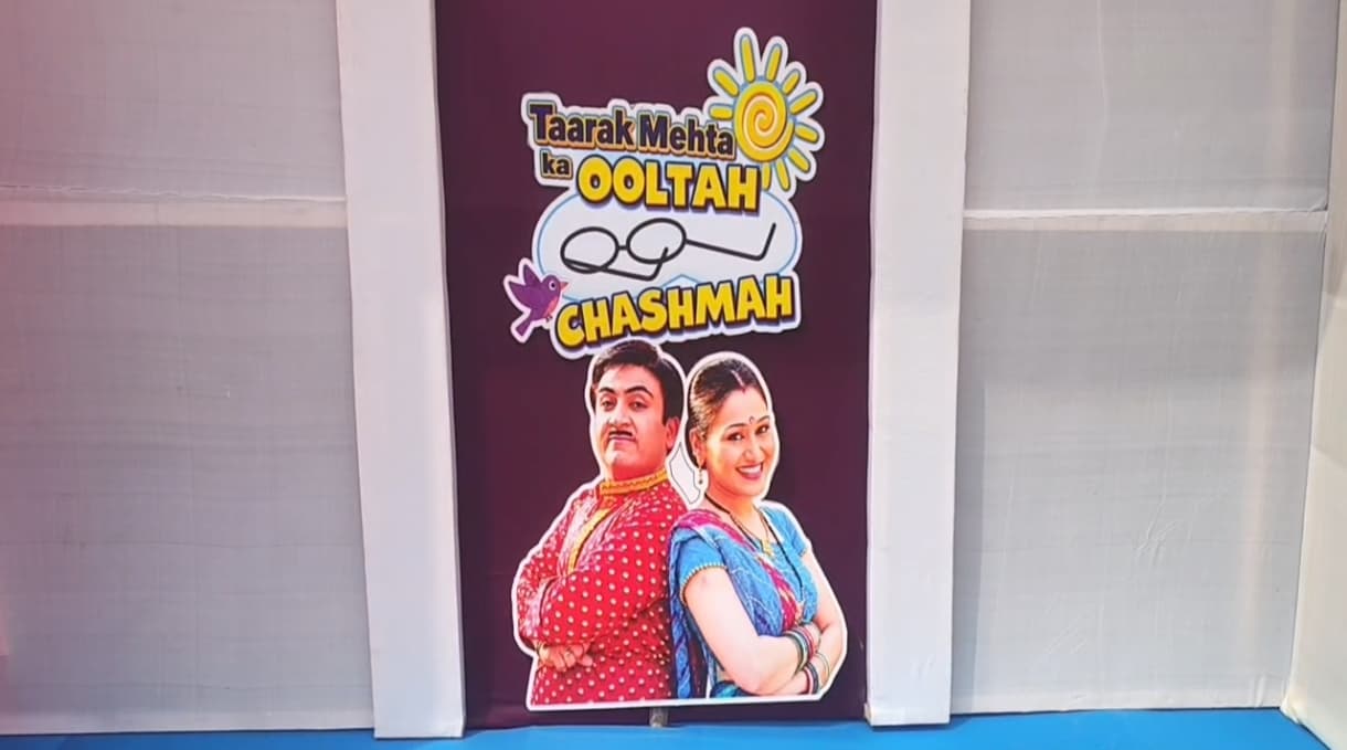 सफेद खंभों और कपड़े की दीवारों के बीच बैंगनी पैनल पर "Taarak Mehta Ka Ooltah Chashmah" का रंगीन लोगो लगा है। उसके नीचे लाल पोल्का-डॉट कुर्ते में एक पुरुष और नीली साड़ी में एक महिला का कटआउट पीठ से पीठ टिकाए खड़ा दिखता है। नीचे नीला फर्श है।