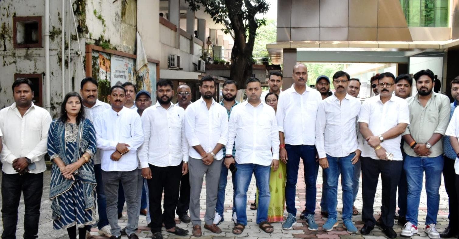 एक इमारत के बाहर बड़ी संख्या में लोग समूह में खड़े दिखाई दे रहे हैं। अधिकांश लोग सफेद और हल्के रंग के कपड़ों में हैं और कैमरे की ओर देख रहे हैं।