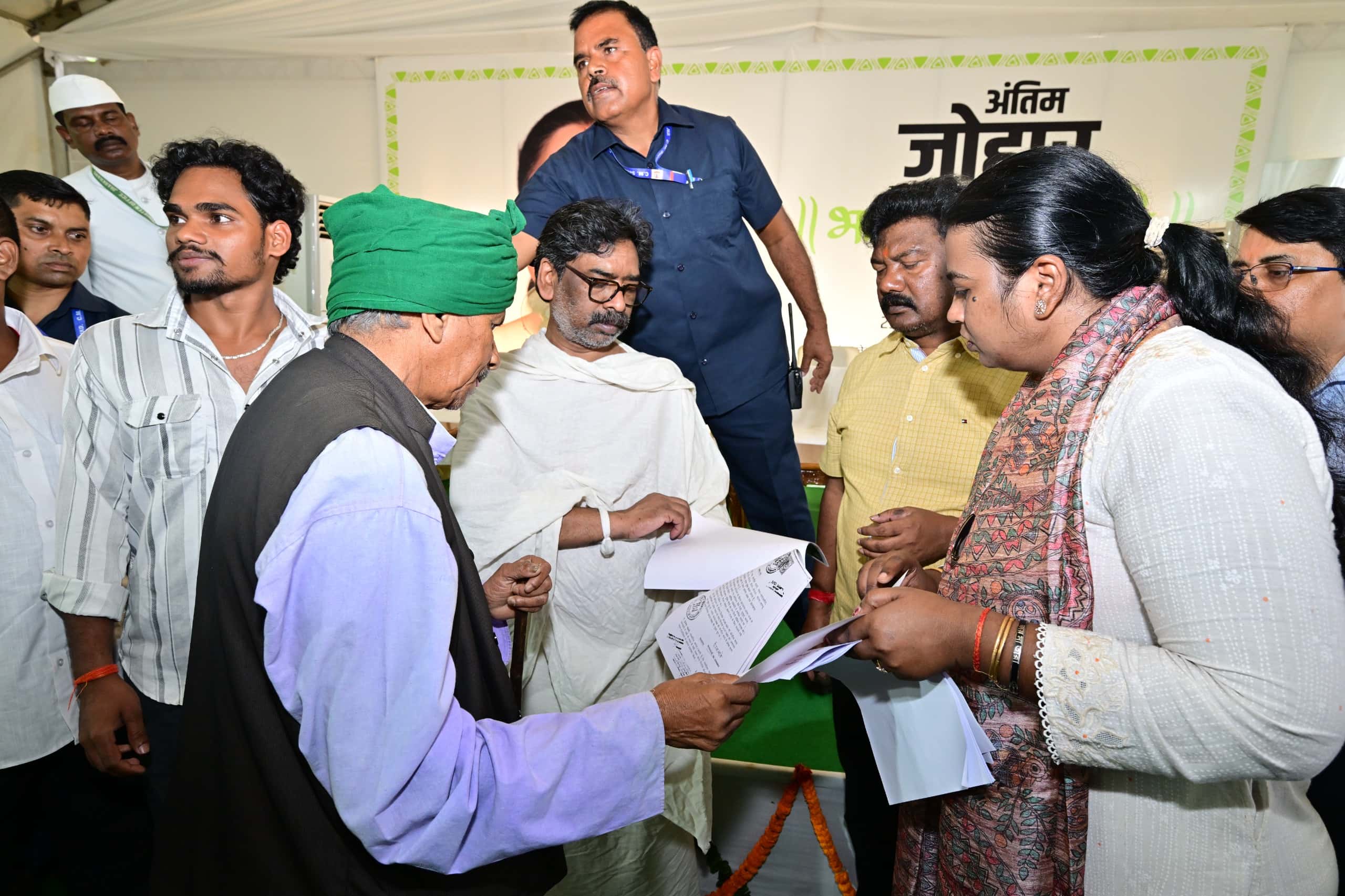 सफेद तंबू के भीतर सफेद चादर ओढ़े एक व्यक्ति कागज देख रहे हैं। उनके सामने हरी पगड़ी और काली जैकेट पहने एक बुजुर्ग तथा प्रिंटेड स्टोल पहने एक महिला भी हाथ में कागज लिए खड़े हैं। पीछे नीली शर्ट वाला एक व्यक्ति वॉकी-टॉकी के साथ खड़ा है और बैनर पर हिंदी में "अंतिम जोहार" का आंशिक पाठ दिखता है।