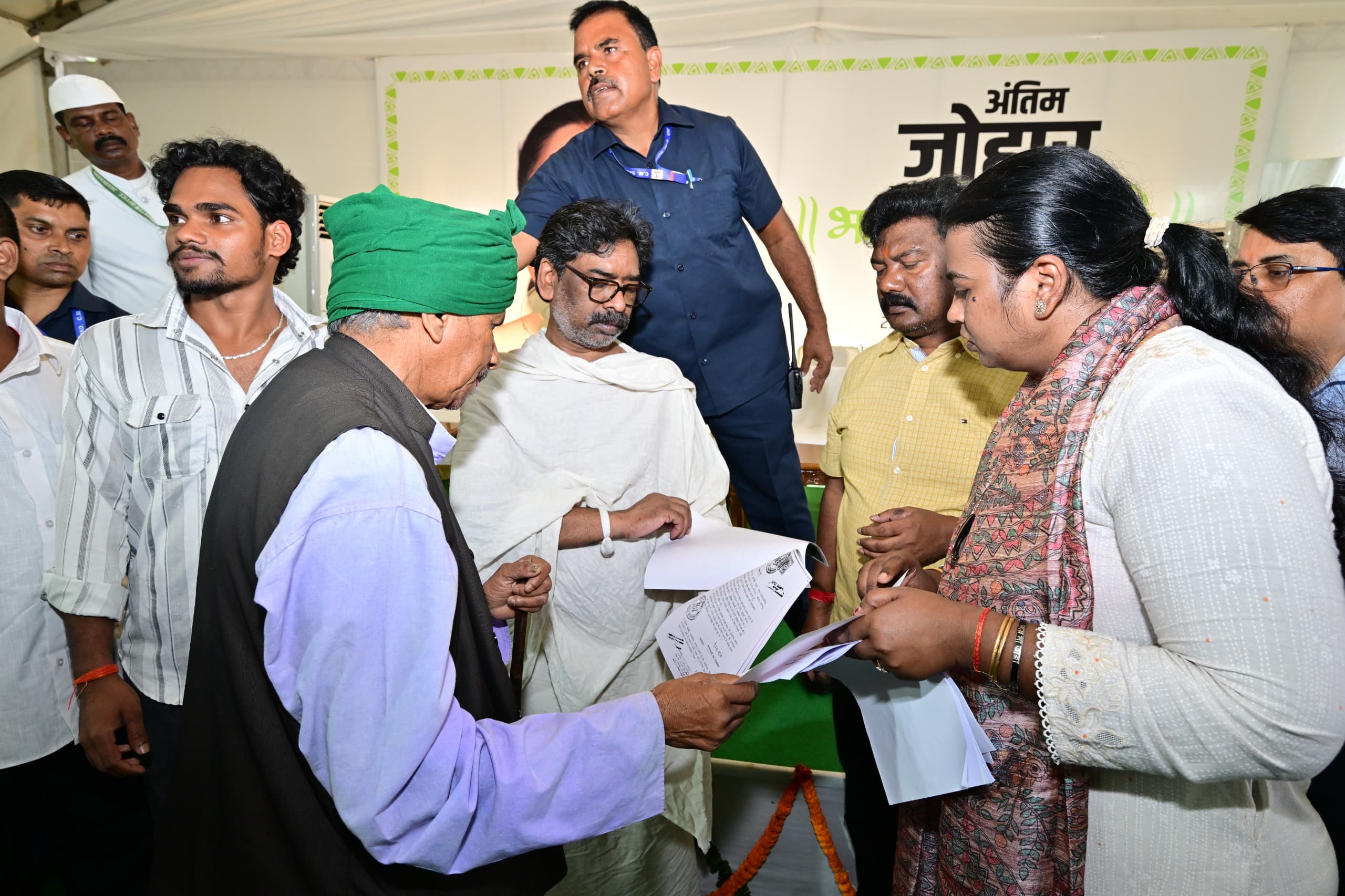 सफेद तंबू के भीतर सफेद चादर ओढ़े एक व्यक्ति कागज देख रहे हैं। उनके सामने हरी पगड़ी और काली जैकेट पहने एक बुजुर्ग तथा प्रिंटेड स्टोल पहने एक महिला भी हाथ में कागज लिए खड़े हैं। पीछे नीली शर्ट वाला एक व्यक्ति वॉकी-टॉकी के साथ खड़ा है और बैनर पर हिंदी में "अंतिम जोहार" का आंशिक पाठ दिखता है।