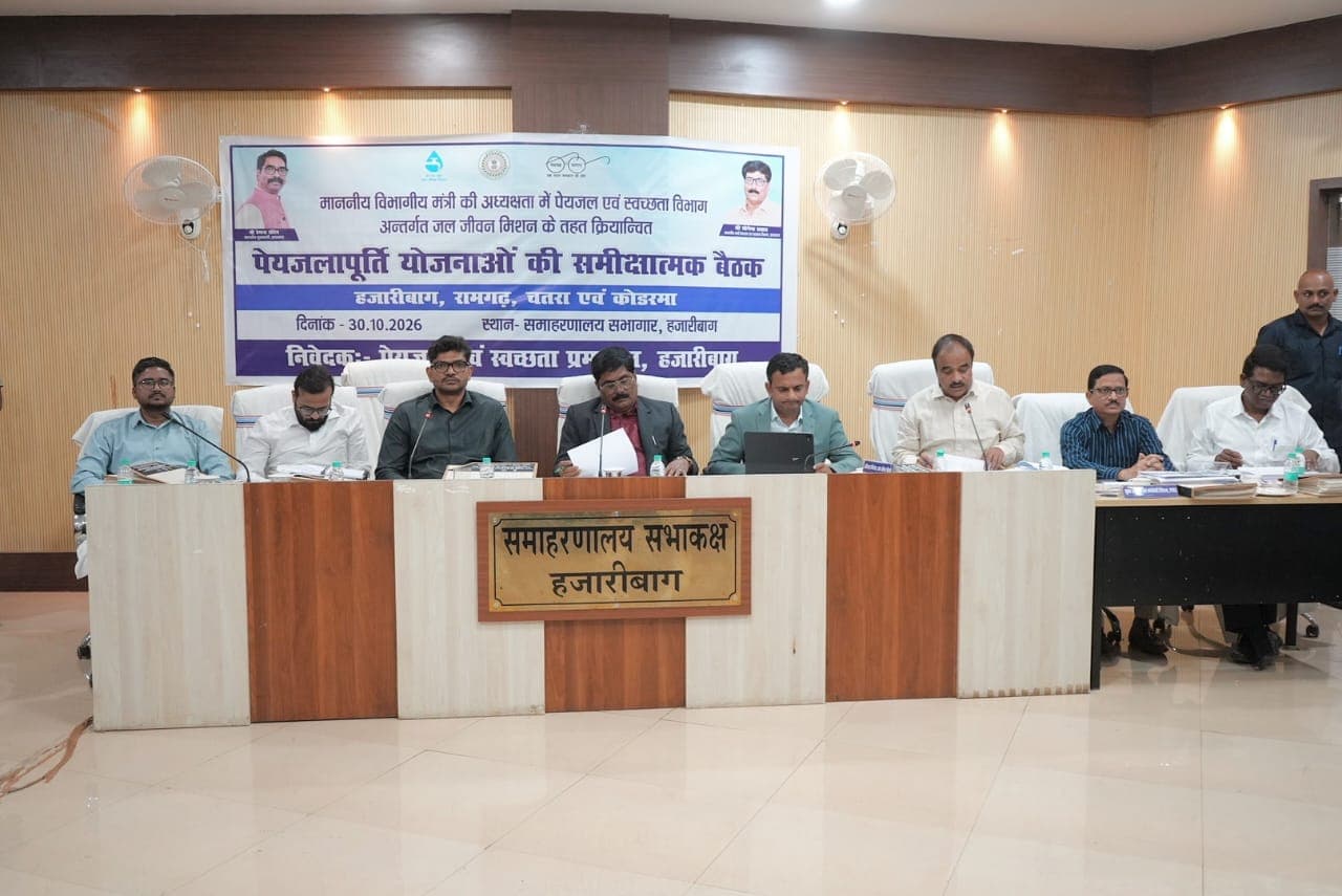 समाहरणालय सभाकक्ष, हजारीबाग में पीछे लगे बैनर के सामने लंबी मेज पर माइक के साथ बैठे कई पुरुष अधिकारी, कुछ के सामने कागजात और एक के सामने लैपटॉप रखा है।