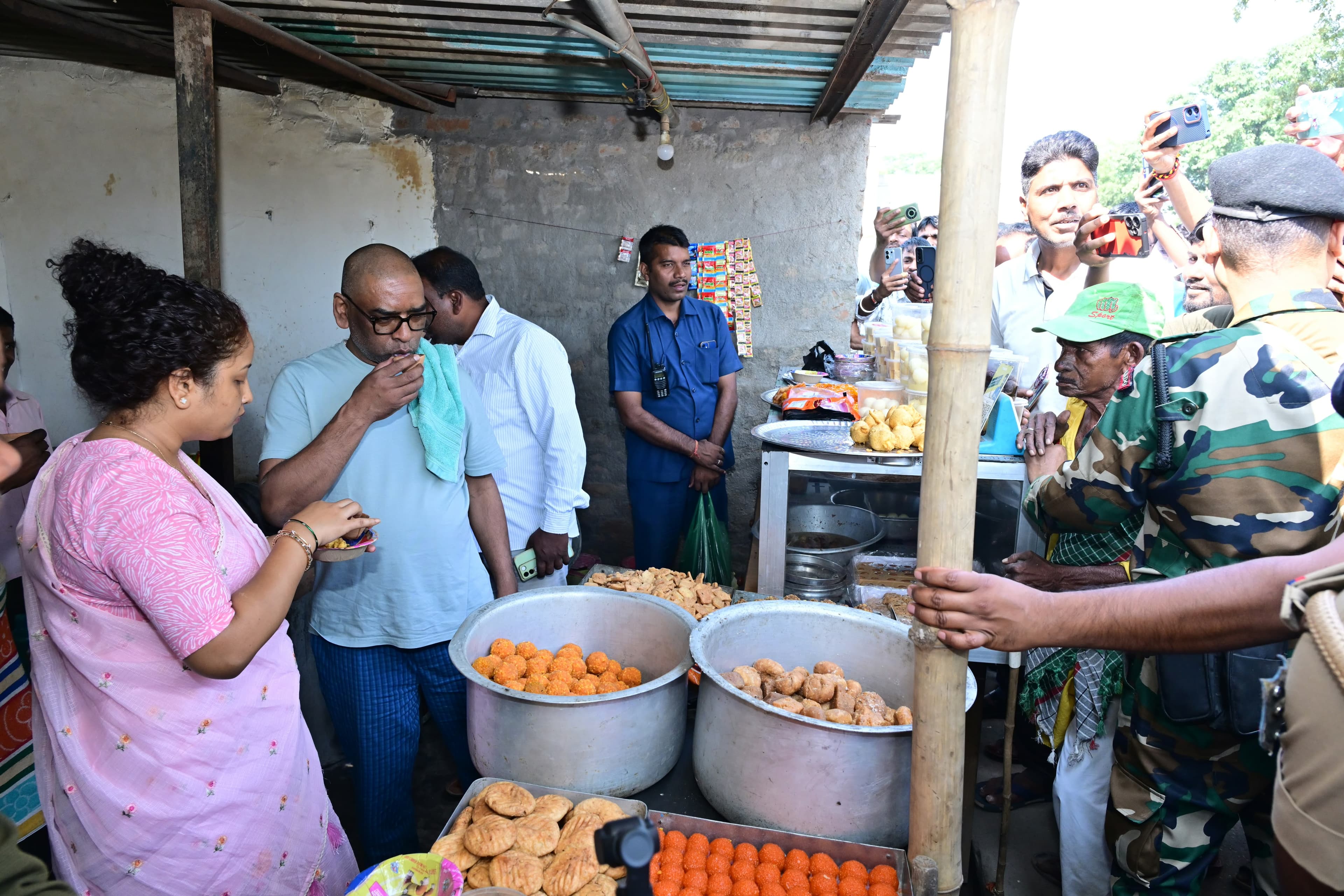 टिन की छत वाली एक छोटी दुकान में बड़े बर्तनों में लड्डू और अन्य मिठाइयां रखी हैं। हल्की नीली टी-शर्ट पहने चश्माधारी एक व्यक्ति हाथ में दोना लेकर कुछ खा रहे हैं, उनके बगल में गुलाबी साड़ी पहने एक महिला भी प्लेट थामे खड़ी हैं। पीछे कैमोफ्लाज वर्दी में सुरक्षाकर्मी और मोबाइल से तस्वीरें लेते कई लोग दिखाई दे रहे हैं।