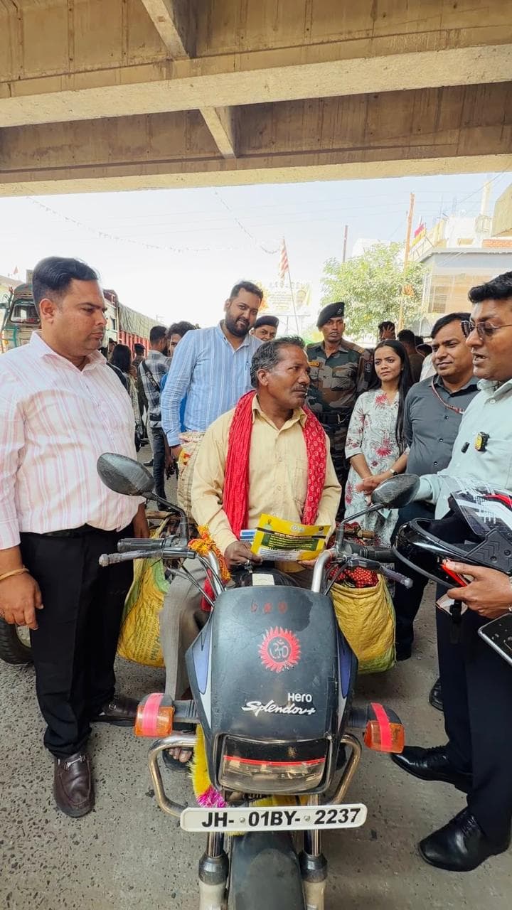 फ्लाईओवर के नीचे एक मोटरसाइकिल पर बैठे व्यक्ति के गले में माला और कंधे पर लाल गमछा है, उनके हाथ में पीला पंपलेट है। आसपास कई पुरुष और एक महिला खड़े हैं, एक व्यक्ति हाथ में काला हेलमेट लिए बात कर रहा है। पीछे वर्दीधारी जवान और ट्रक दिख रहे हैं।