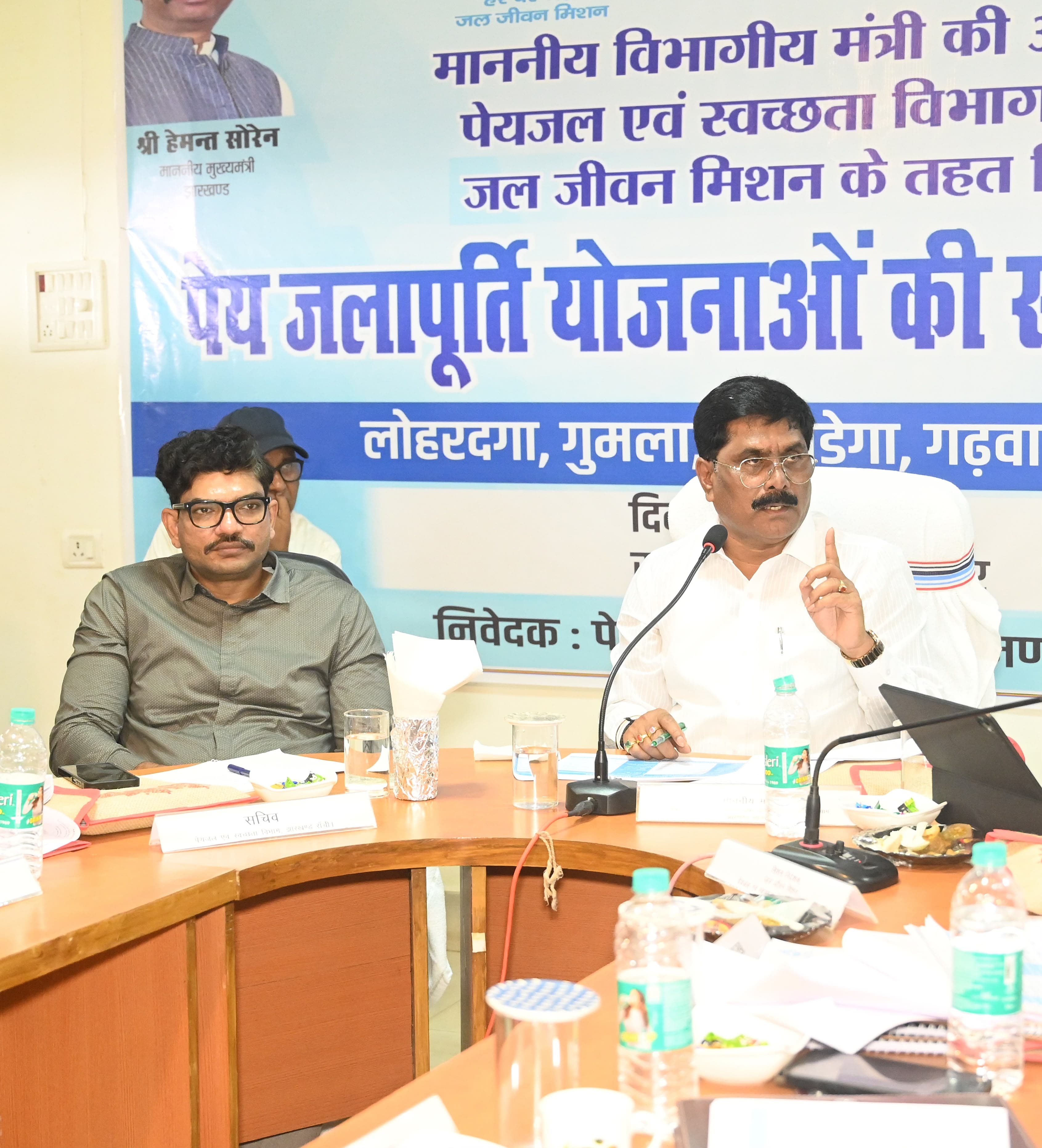 सफेद कुर्ते में एक व्यक्ति माइक के सामने उंगली उठाकर बोलते हुए, उनके बगल में चश्मा लगाए धूसर शर्ट वाला व्यक्ति बैठा है। मेज पर सचिव लिखी नामपट्टिका रखी है।