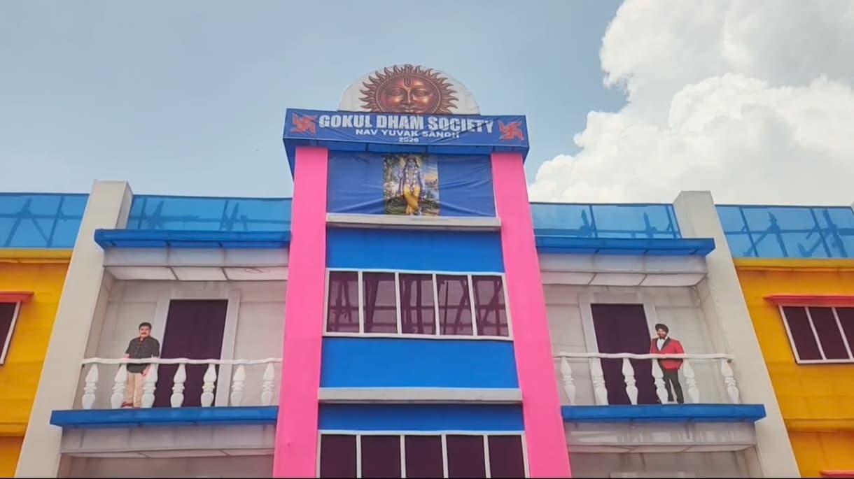 गुलाबी, नीले और पीले रंगों में सजी दो मंजिला इमारत जैसा पंडाल दिख रहा है। ऊपर लगे नीले बैनर पर अंग्रेजी में "GOKUL DHAM SOCIETY, NAV YUVAK SANGH, 2026" लिखा है और उसके ऊपर सूर्य की आकृति बनी है। बैनर के नीचे एक देवता की तस्वीर लगी है। दोनों ओर की बालकनी में एक-एक व्यक्ति का कटआउट खड़ा है, जिनमें से एक ने लाल ब्लेजर और पगड़ी पहनी है।