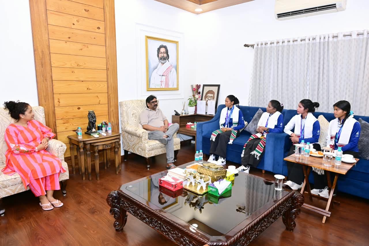 एक कमरे में नीले सोफे पर नीली जैकेट और सफेद अंगवस्त्र पहने चार महिलाएं बैठी हैं। सामने कुर्सी पर एक पुरुष बैठे हैं और बाईं ओर गुलाबी-नारंगी धारीदार पोशाक में एक महिला हैं। पीछे दीवार पर एक व्यक्ति का फ्रेम में लगा चित्र दिखता है।