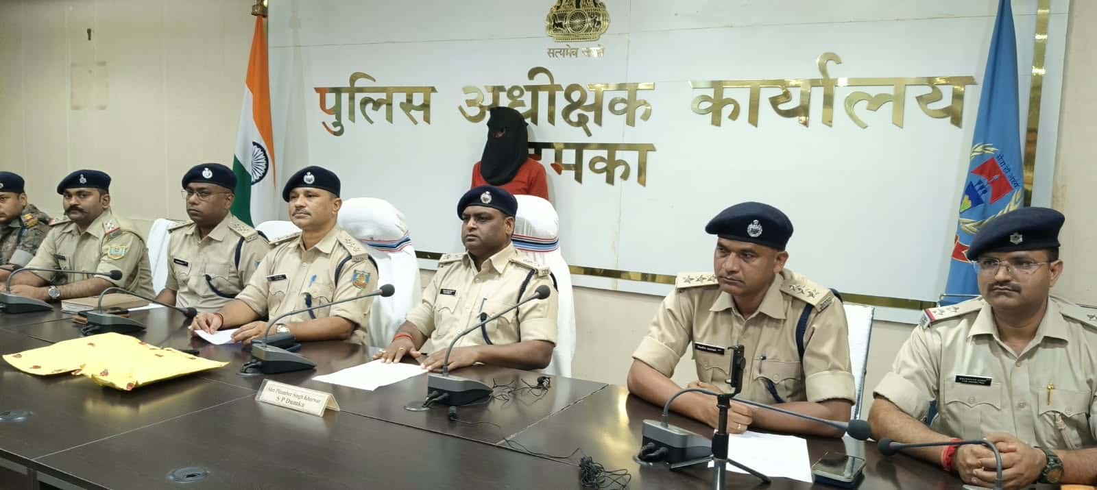 पुलिस अधीक्षक कार्यालय के बोर्ड के सामने मेज के पीछे खाकी वर्दी और नीली टोपी पहने कई पुलिसकर्मी बैठे हैं। पीछे काले कपड़े से चेहरा ढके, लाल कपड़े पहने एक व्यक्ति बैठा है। मेज पर माइक और एक पीला पैकेट रखा है।