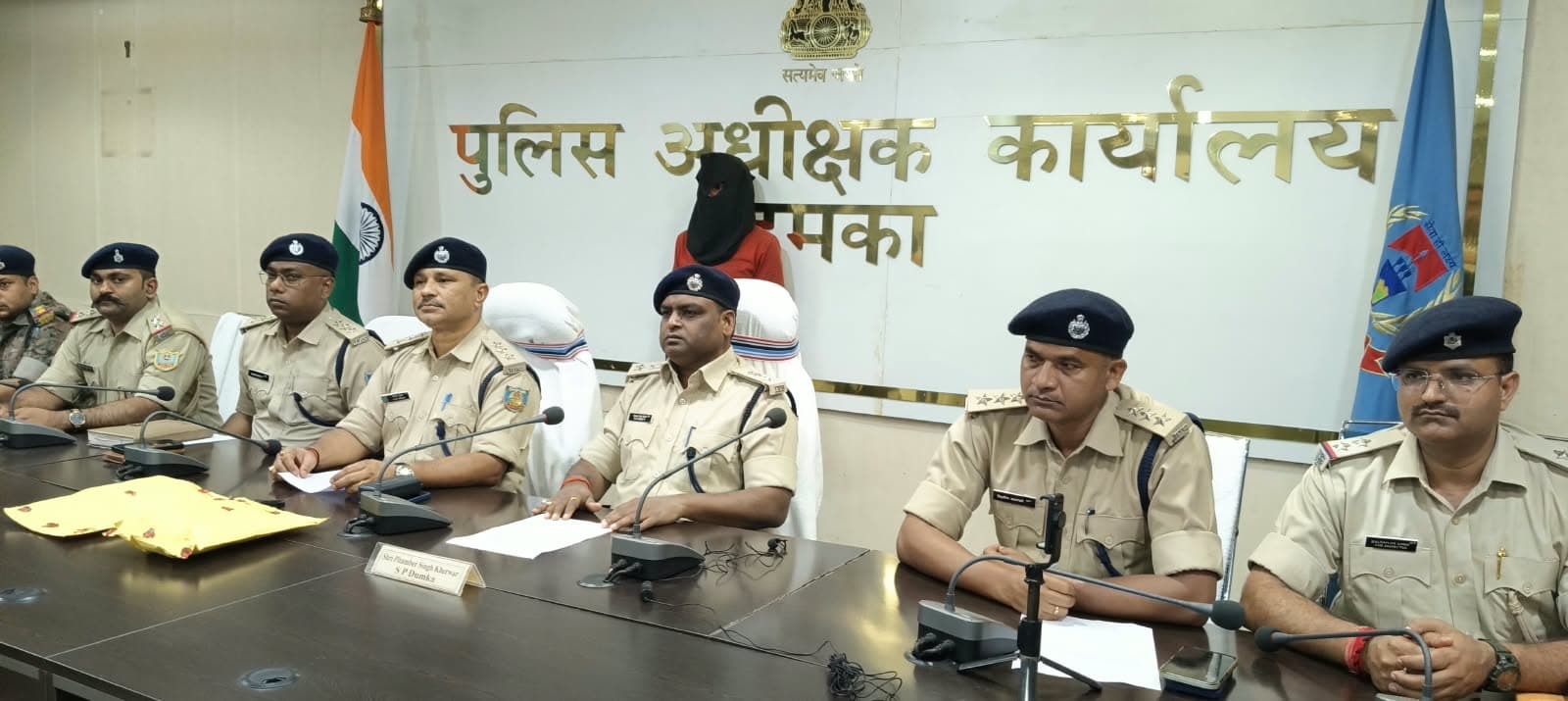 पुलिस अधीक्षक कार्यालय के बोर्ड के सामने मेज के पीछे खाकी वर्दी और नीली टोपी पहने कई पुलिसकर्मी बैठे हैं। पीछे काले कपड़े से चेहरा ढके, लाल कपड़े पहने एक व्यक्ति बैठा है। मेज पर माइक और एक पीला पैकेट रखा है।
