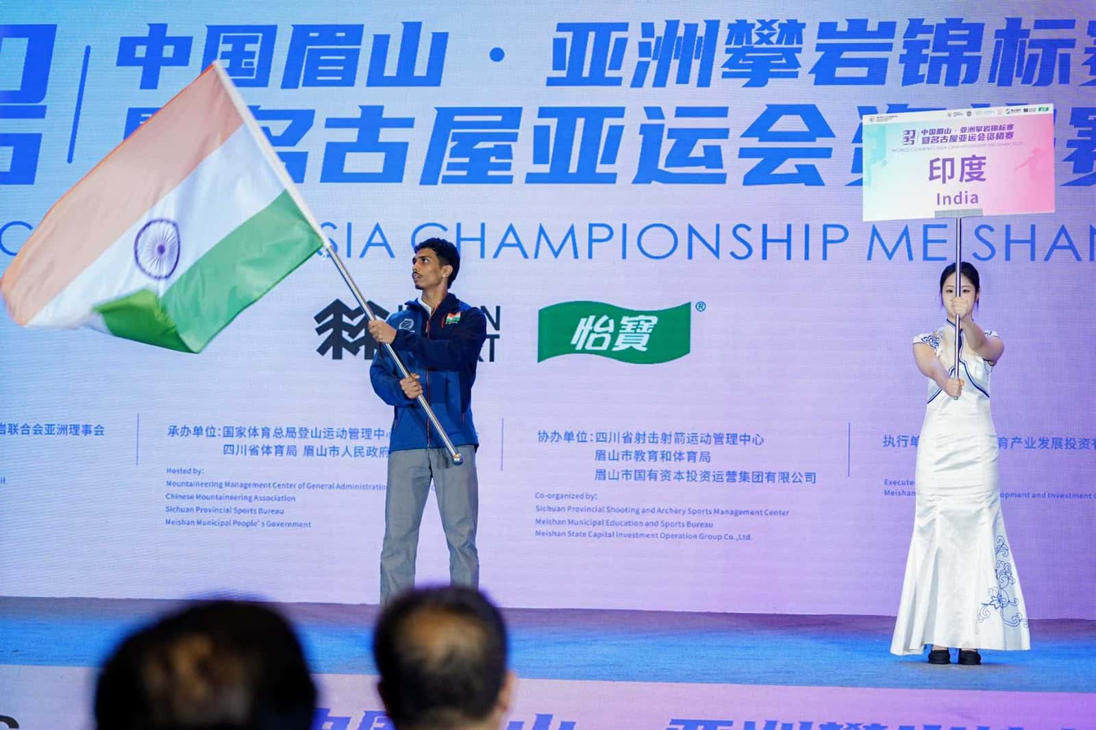 चीन में आयोजित क्लाइम्बिंग प्रतियोगिता के एक समारोह के दौरान भारतीय दल का एक सदस्य तिरंगा लेकर मंच पर नजर आ रहा है। मंच पर भारत का बोर्ड लगा है और पीछे प्रतियोगिता से संबंधित बैनर दिखाई दे रहा है।