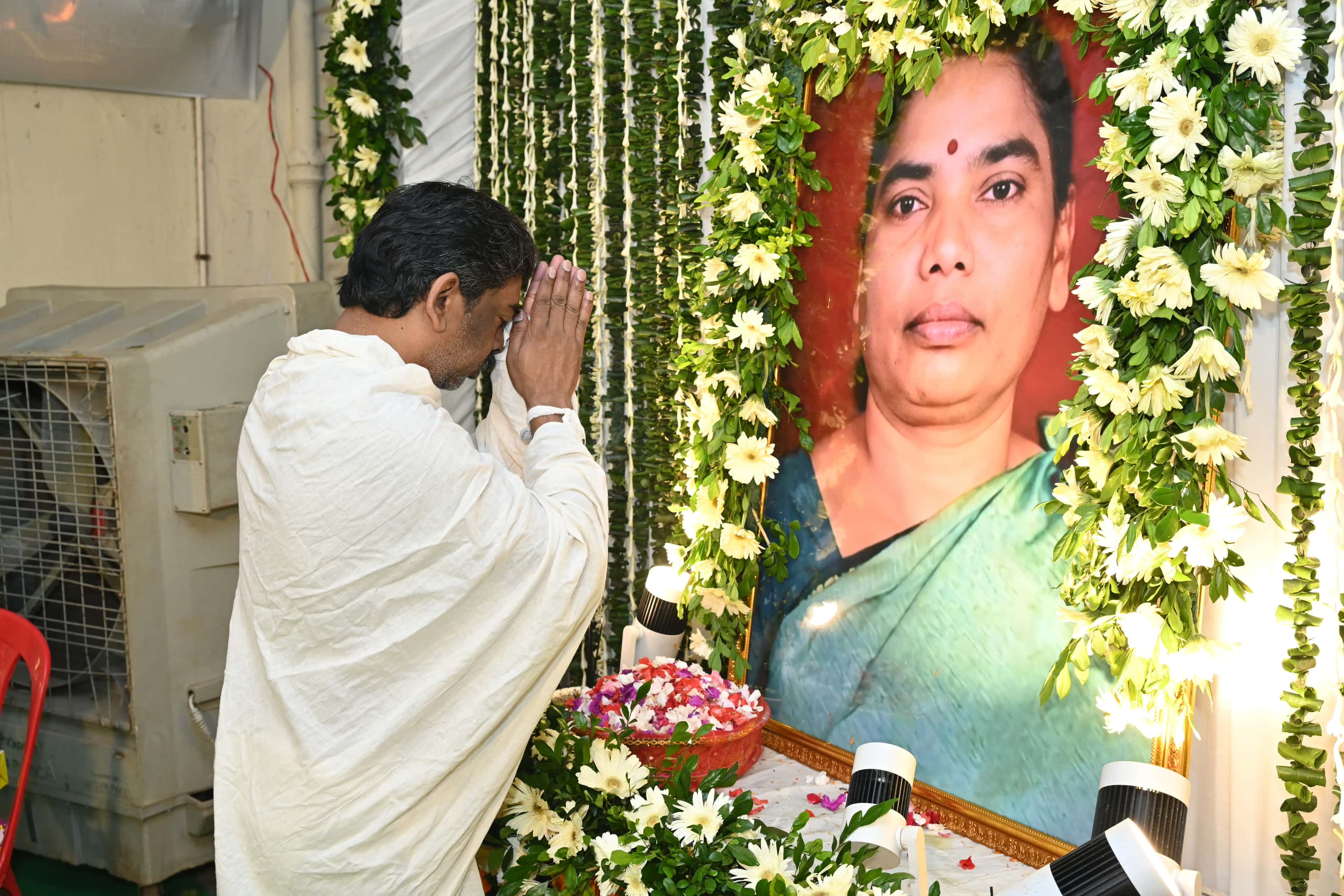सफेद चादर ओढ़े एक पुरुष हाथ जोड़कर सिर झुकाए खड़े हैं। उनके सामने फूलों और हरी पत्तियों की लड़ियों से सजी एक महिला की बड़ी तस्वीर रखी है, जिसके नीचे फूलों की पंखुड़ियों से भरी थाली है।